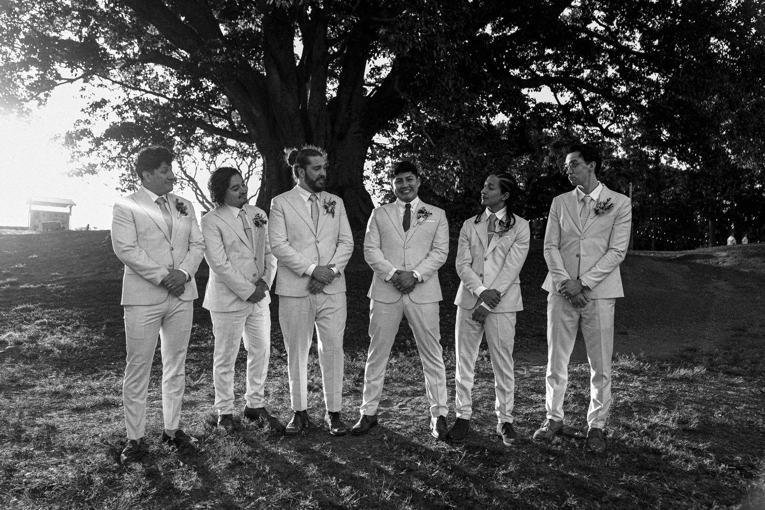 A group of people dressed in white cricket uniforms stand in a row outdoors for a team photograph.