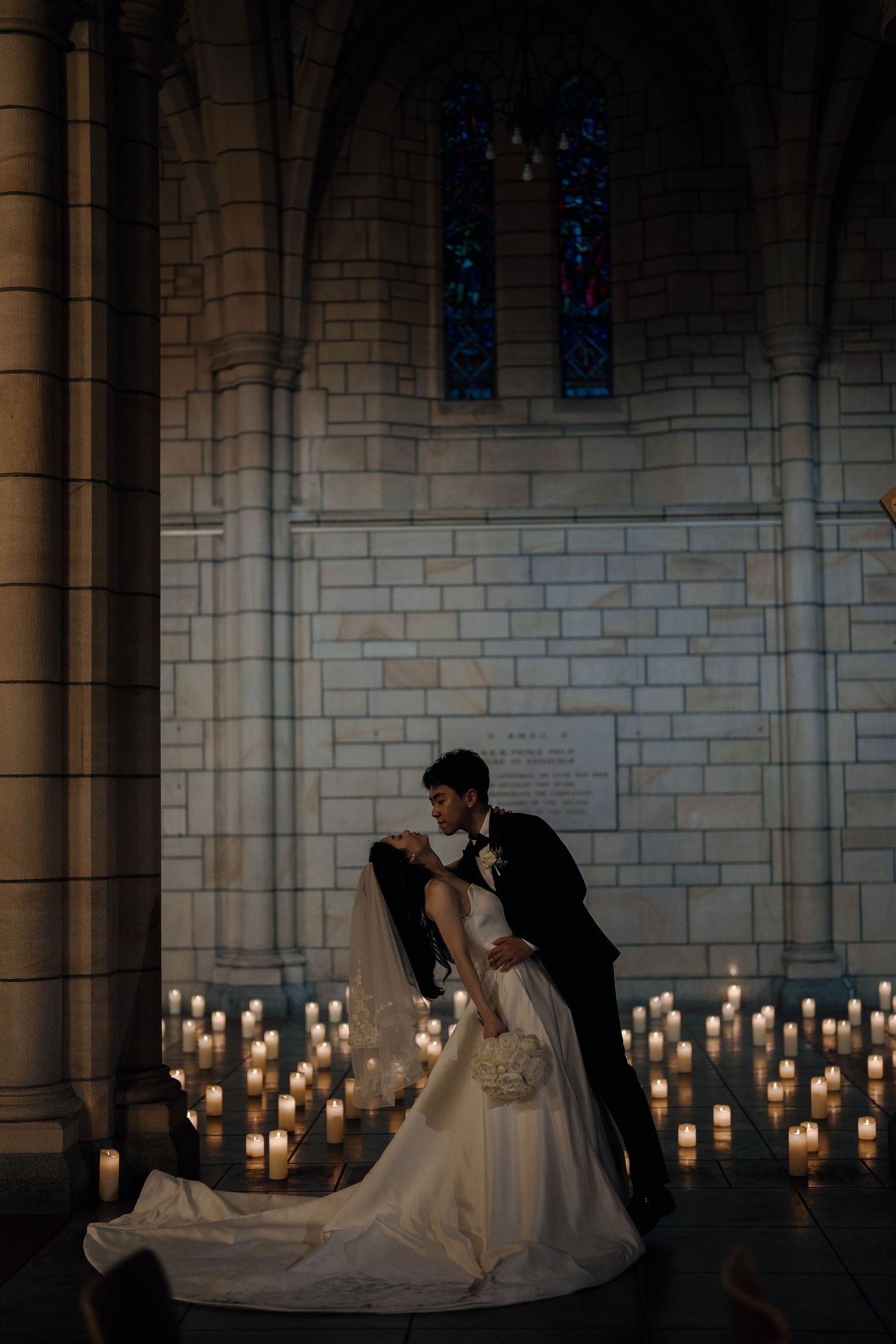 A romantic couple in wedding attire dances in a candlelit church with stone walls and stained glass windows.