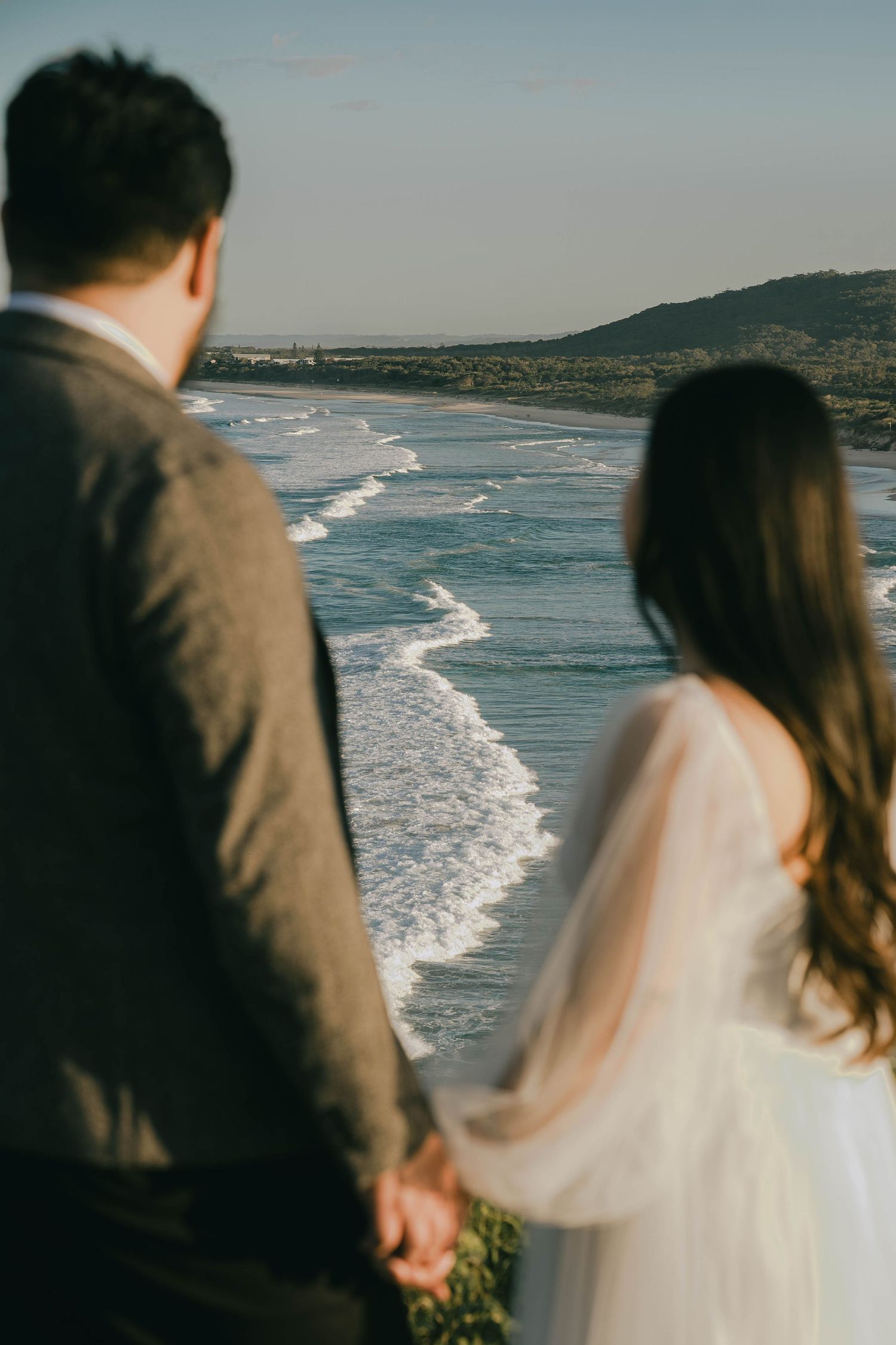 A couple in wedding attire stands at a coastal overlook watching waves crash on a scenic beach below.