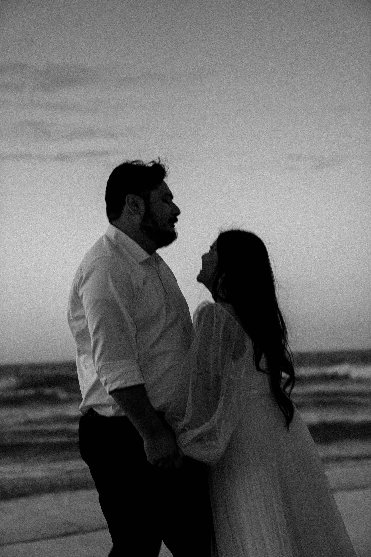 A romantic black and white silhouette of a couple embracing on the beach at dusk with ocean waves in the background.