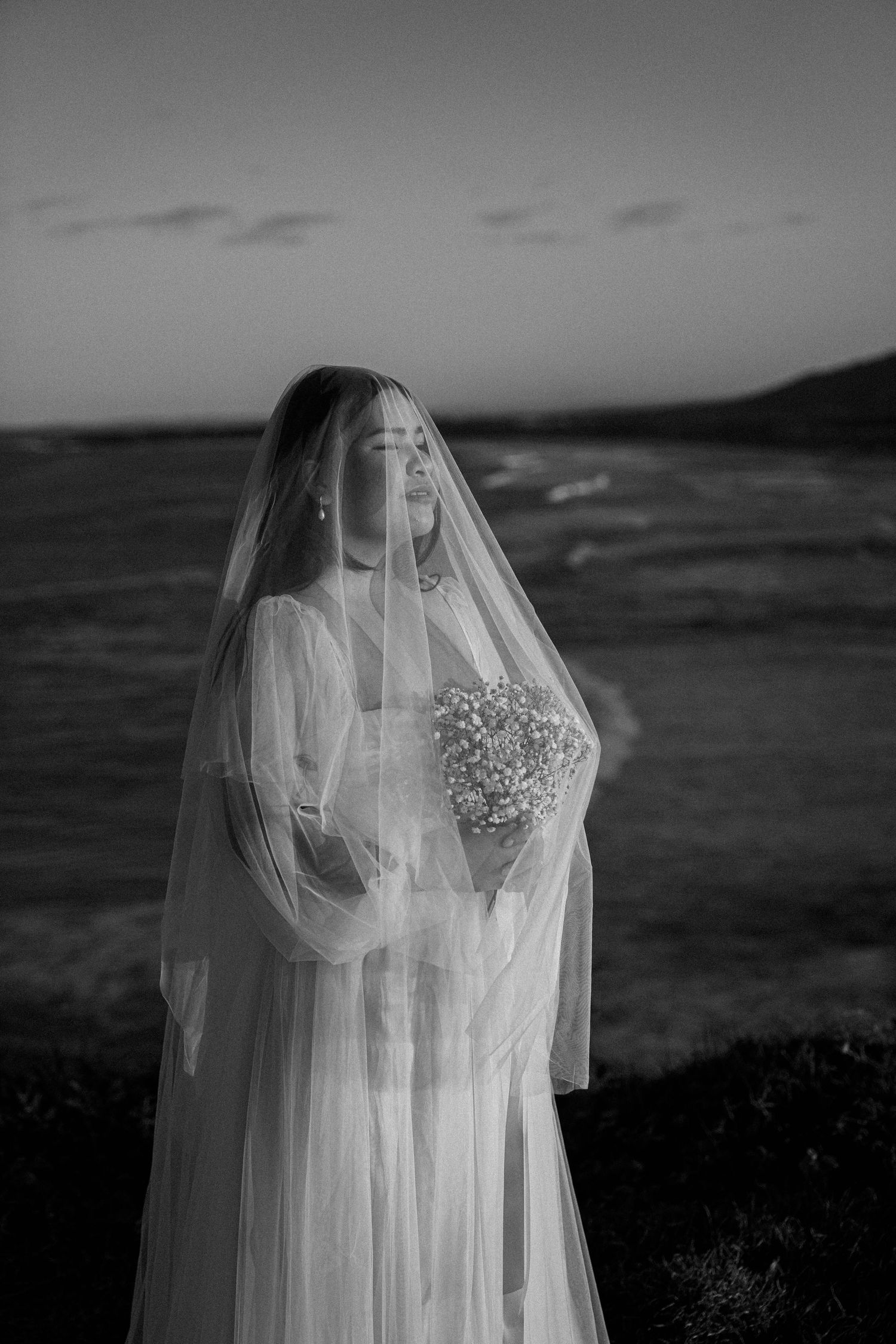 A bride in a flowing white veil and gown holds a bouquet against a moody black and white landscape at dusk.