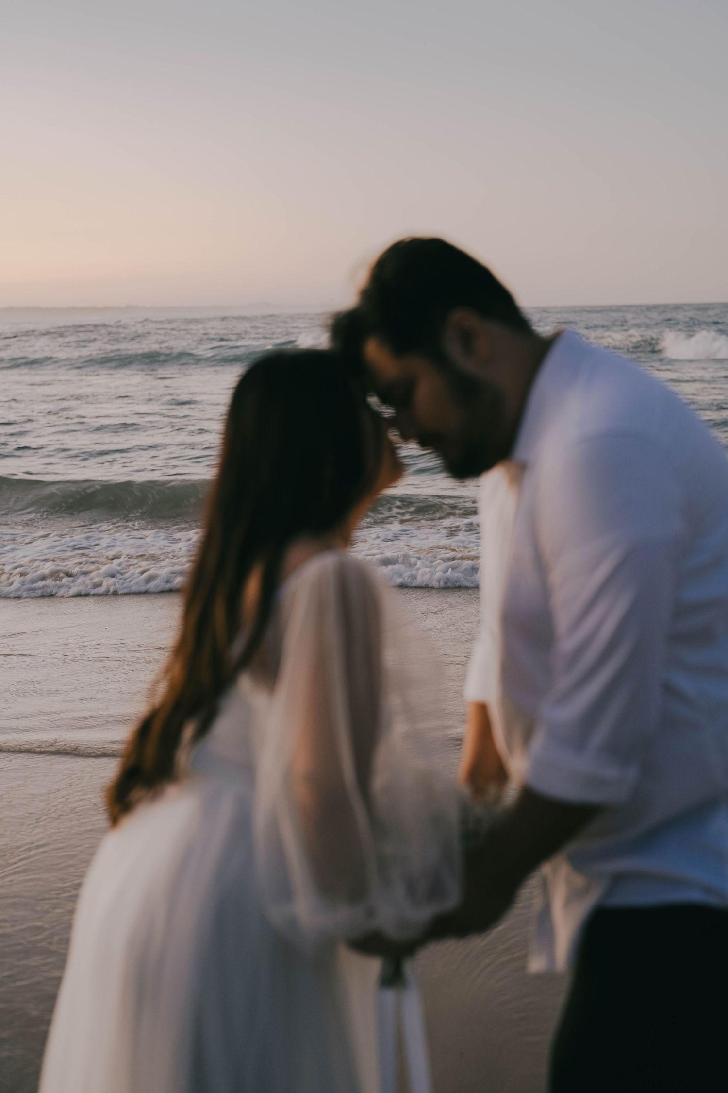 A romantic couple embraces on a beach at sunset with ocean waves in the background.
