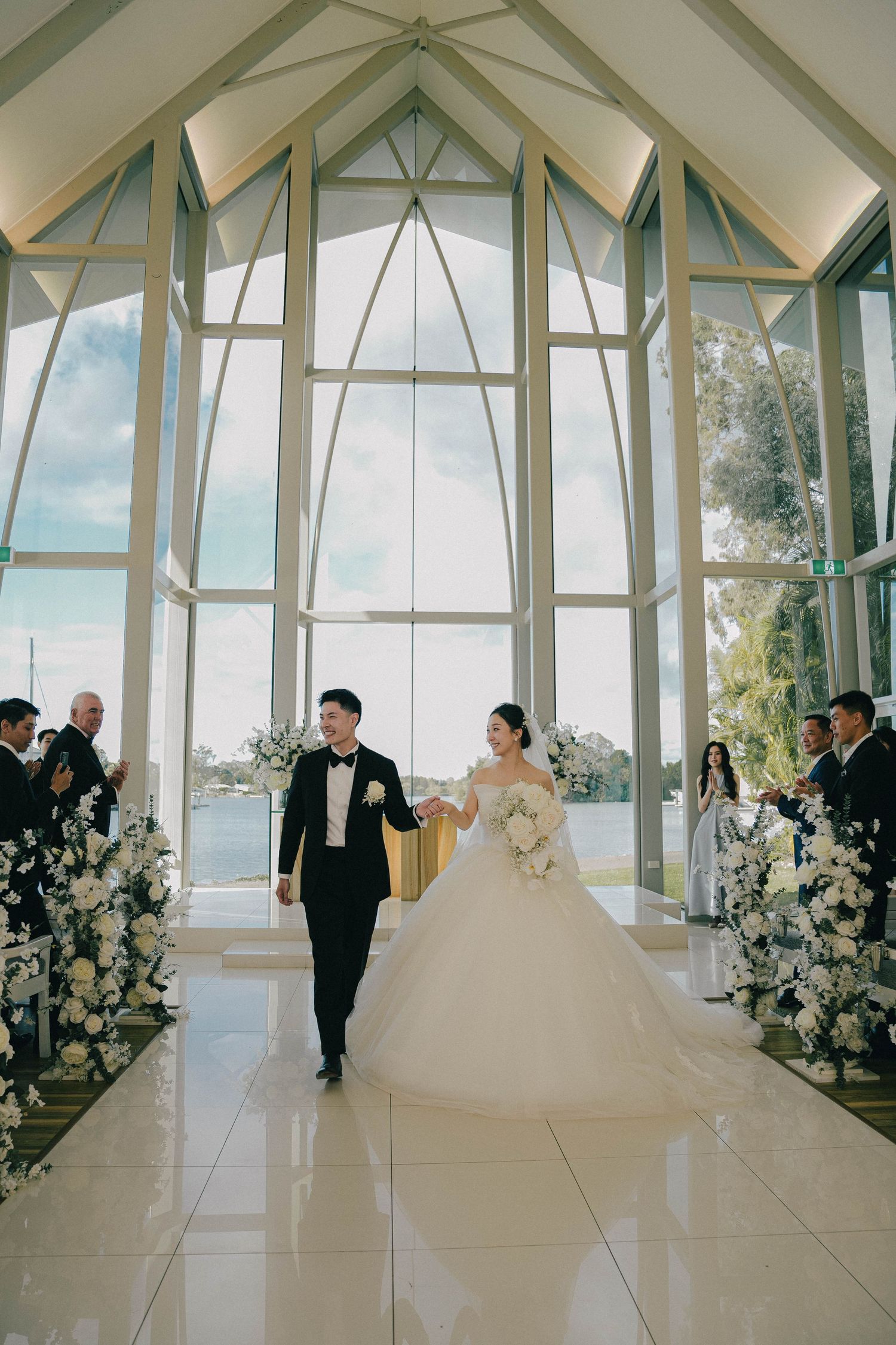 A bride in a flowing white gown walks down a modern glass chapel aisle adorned with white floral arrangements.