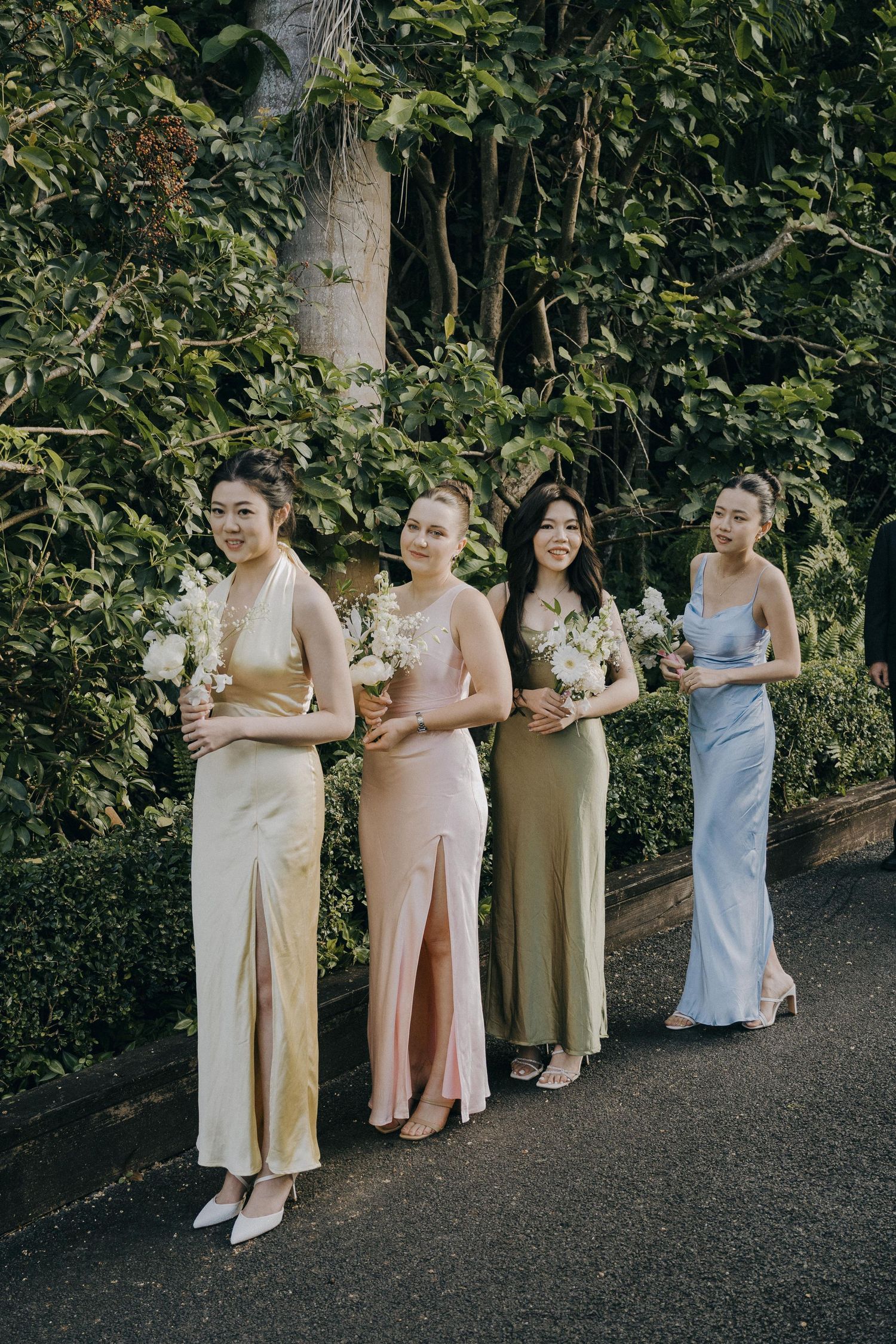 Four bridesmaids wearing elegant silk gowns in neutral tones stand together holding bouquets on a garden path.