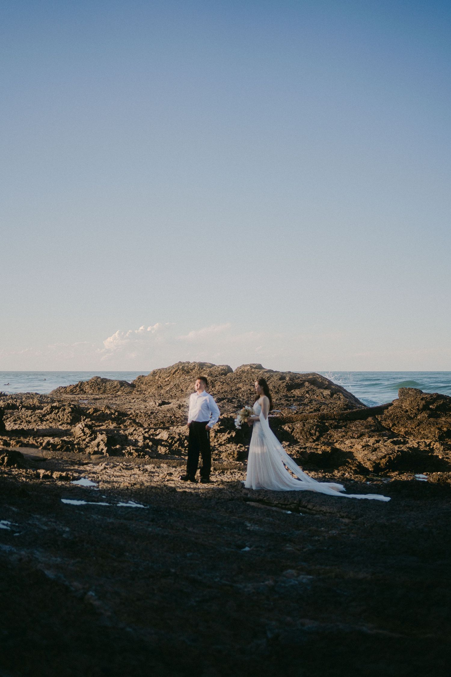 A couple in wedding attire stands on dramatic rocky coastal cliffs overlooking the ocean at sunset.
