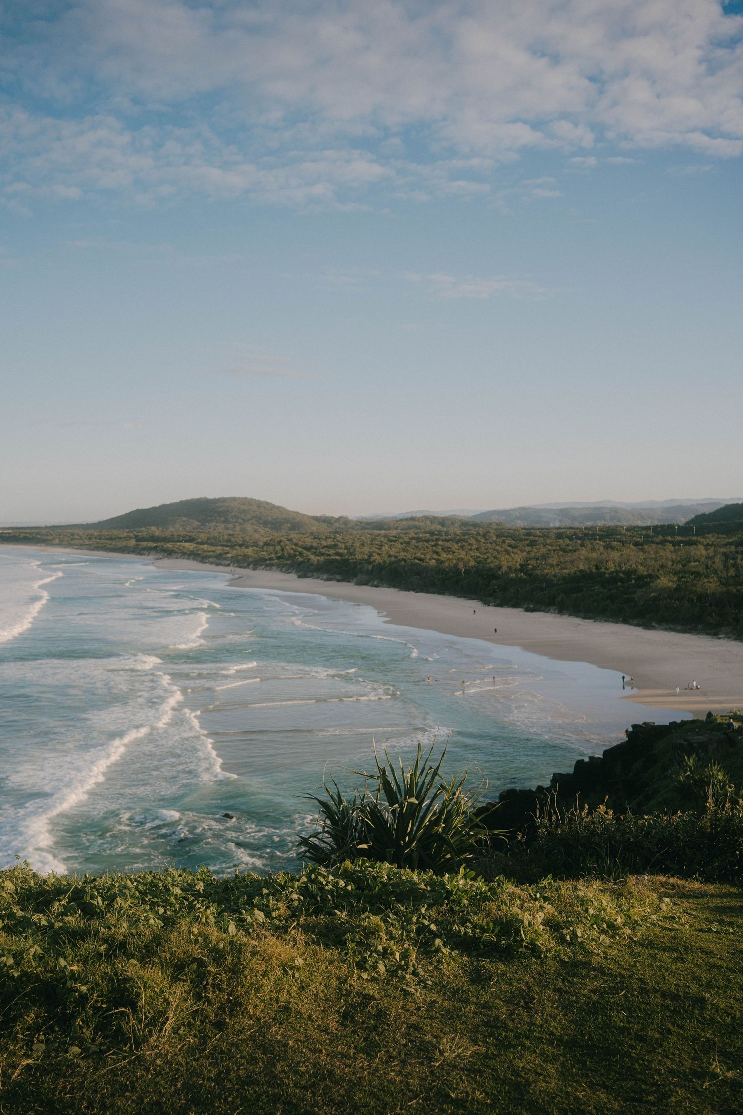 Panoramic coastal view showing rolling waves meeting a curved sandy beach with green hills and vegetation in the foreground.