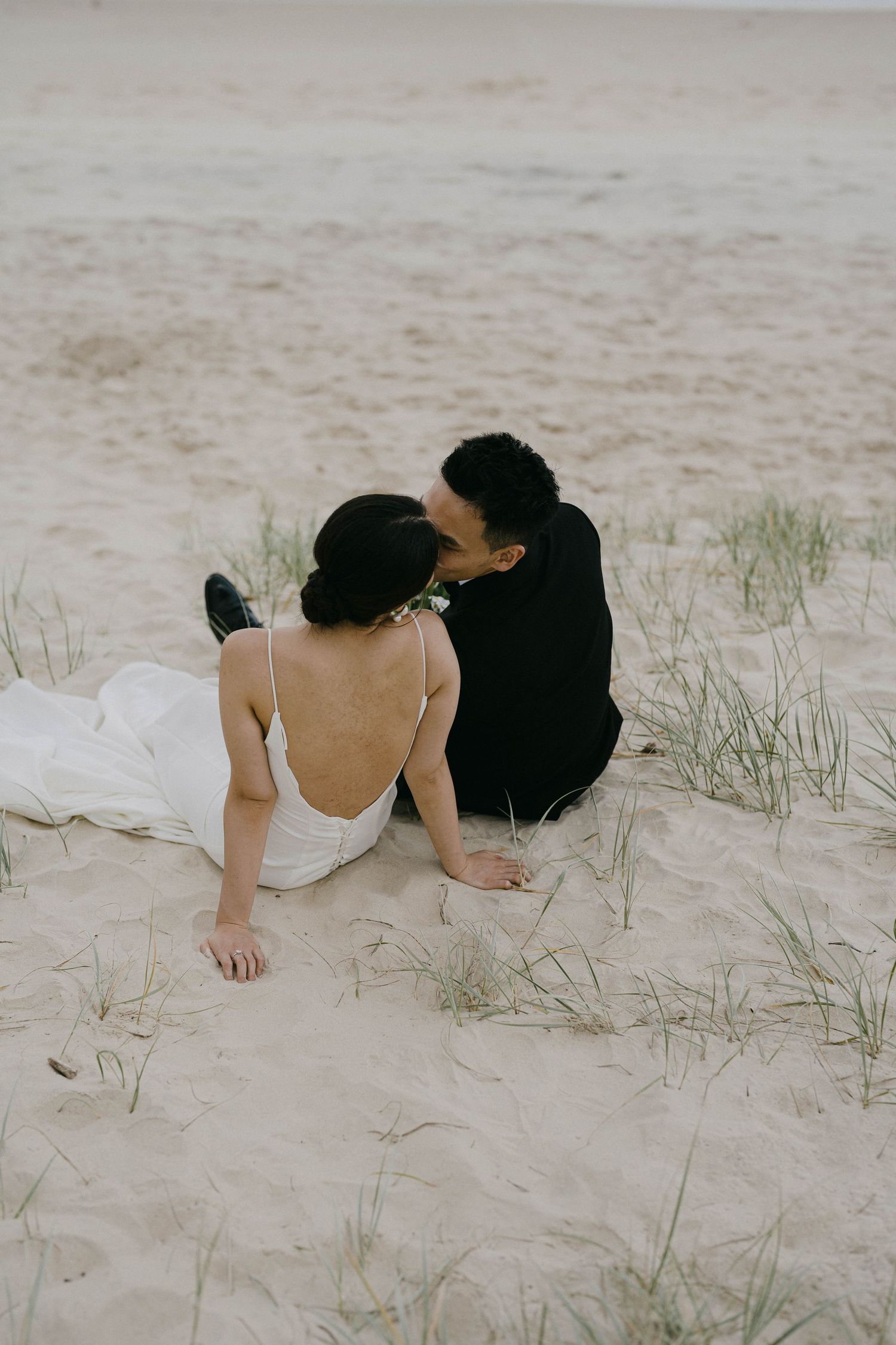 A couple in white clothing lies together on sandy beach dunes with sparse grass during a romantic moment.