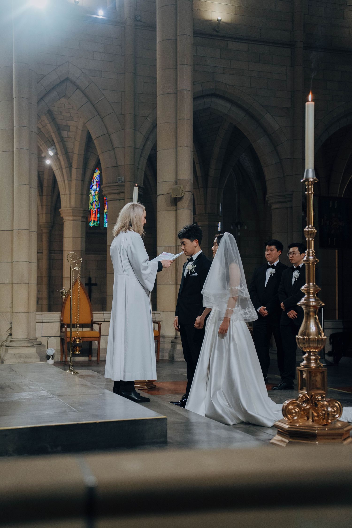A wedding ceremony takes place inside a grand cathedral with gothic stone arches and ornate golden candlesticks.