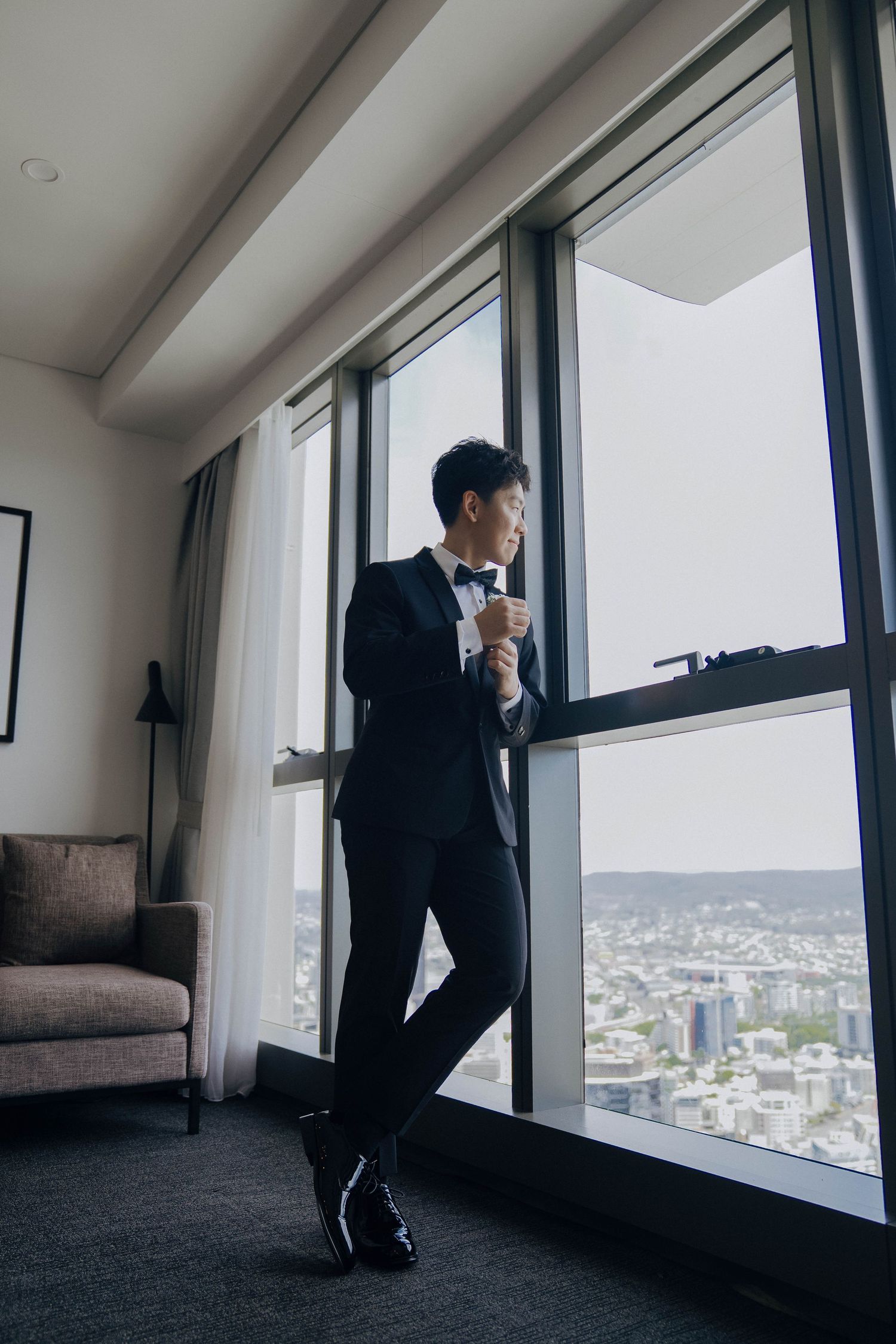 A person in a dark suit sits contemplatively by large windows overlooking a cityscape view.