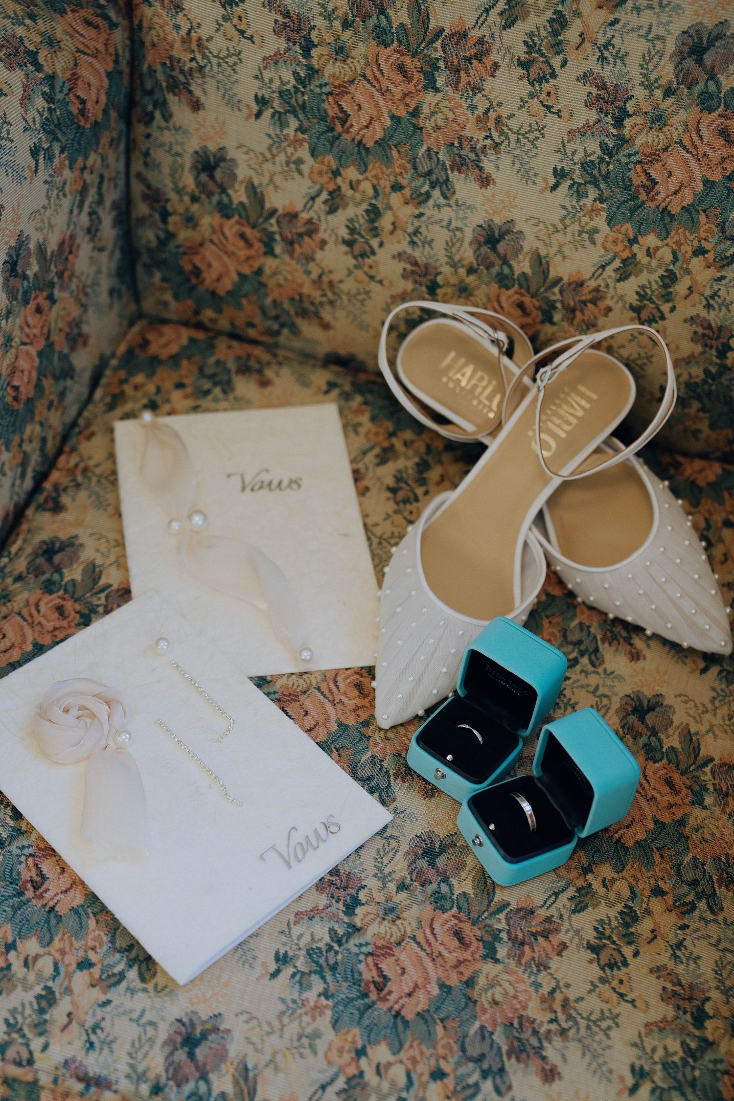 Wedding details including white lace shoes, vows on paper, and a turquoise ring box arranged on a floral patterned surface.