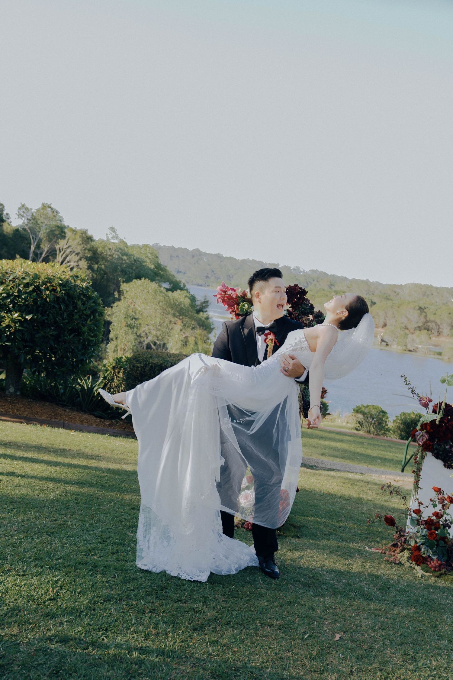 A couple in wedding attire share a romantic dip pose on a grassy hillside with floral decorations and mountains in background.