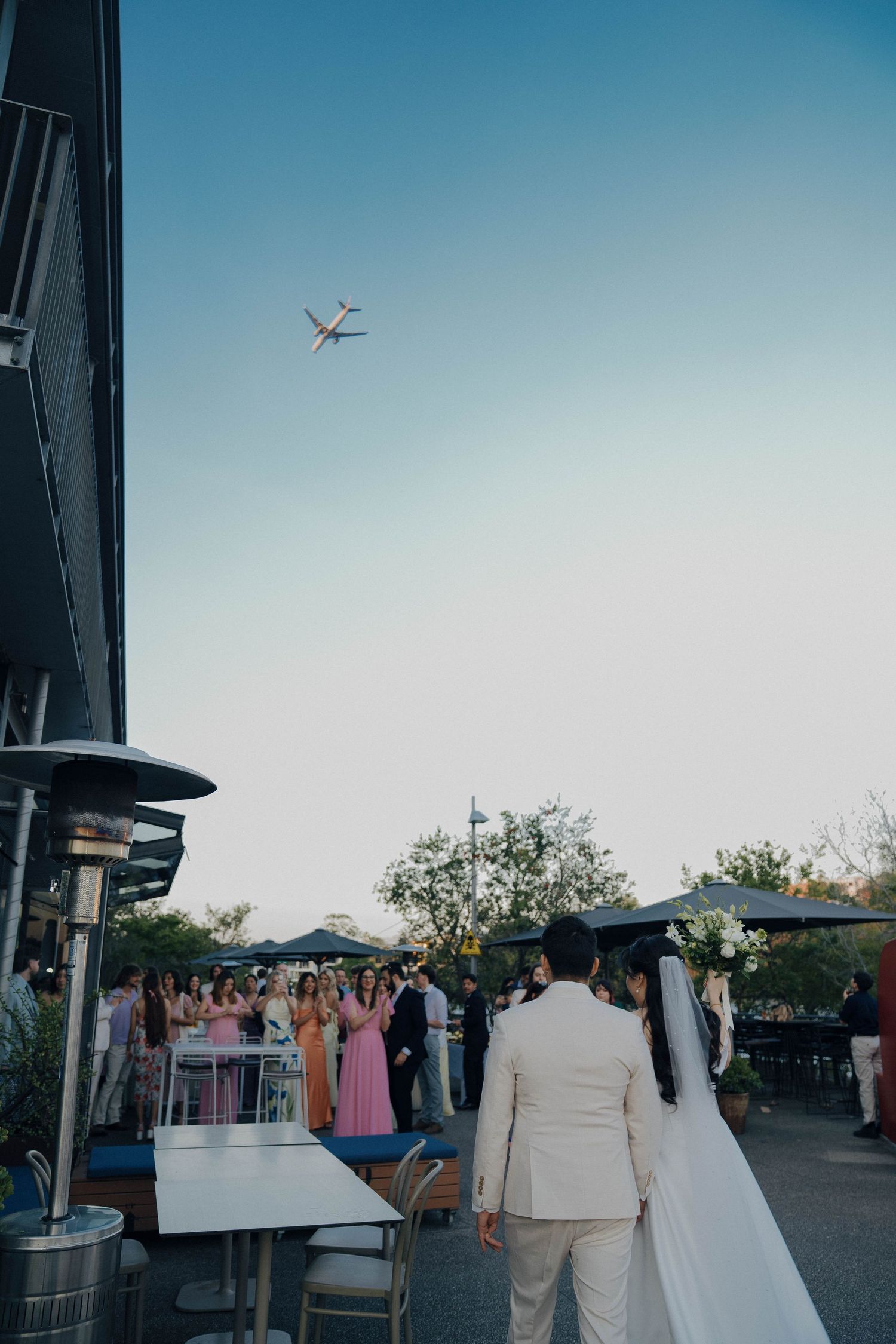 A plane flies overhead during an outdoor wedding reception as guests gather on a patio at dusk.