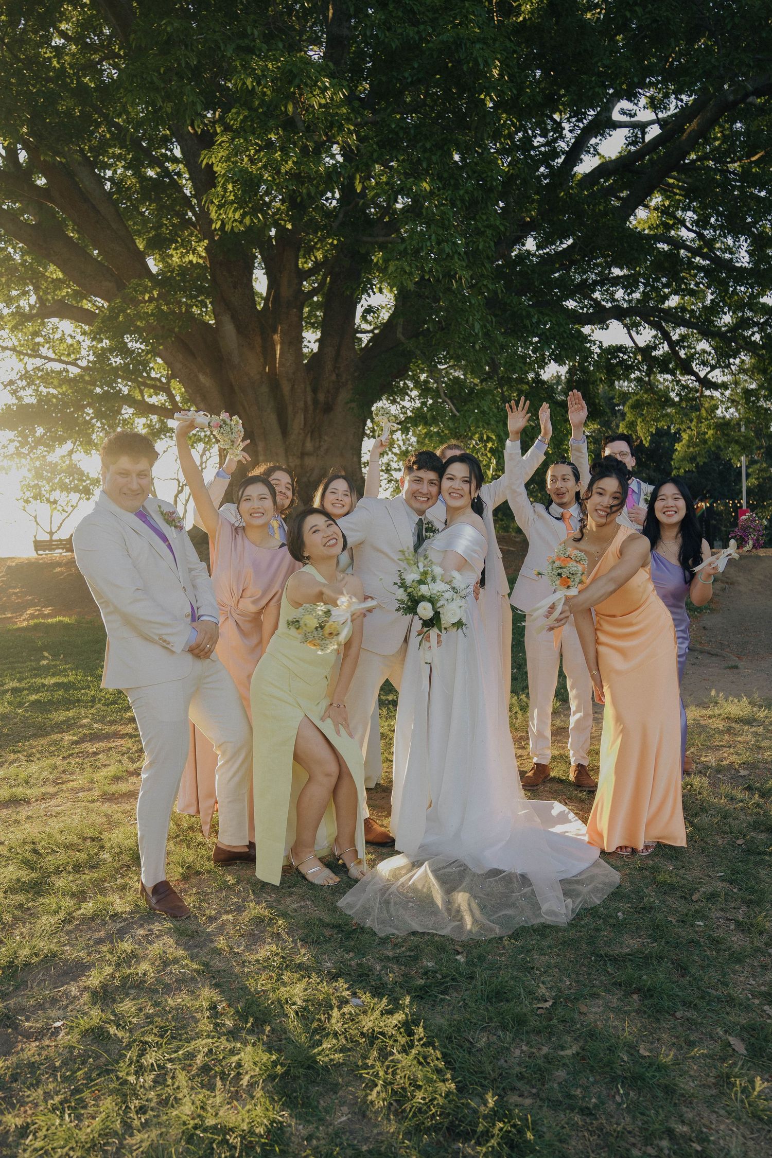 Wedding party in soft pastel colors poses together on grassy hillside during golden hour sunset.