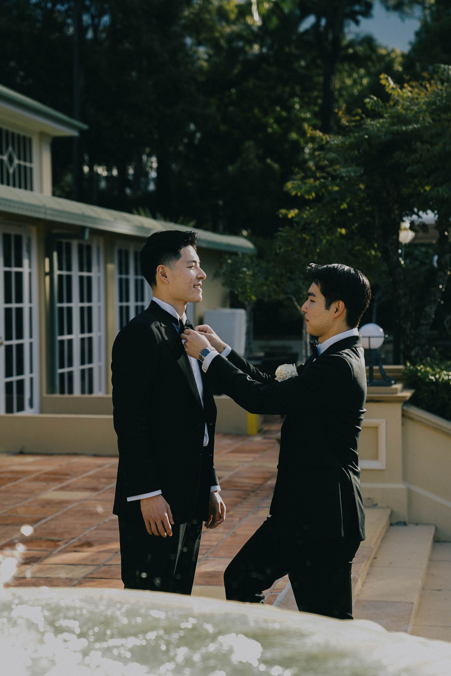 Two people in black suits interacting on a paved terrace outside a white building with windows.