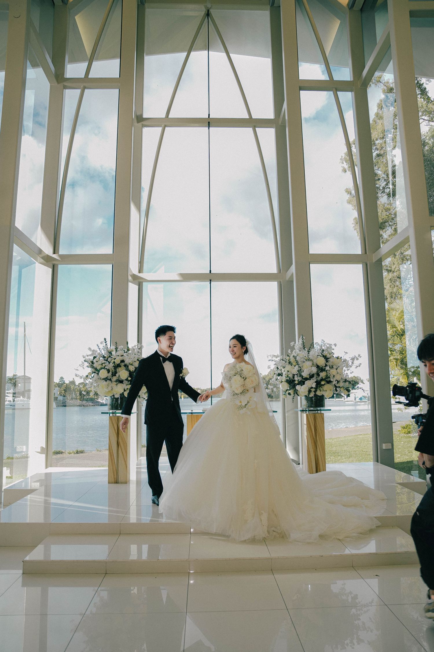 A couple in wedding attire holds hands inside a modern chapel with floor-to-ceiling windows and floral arrangements.