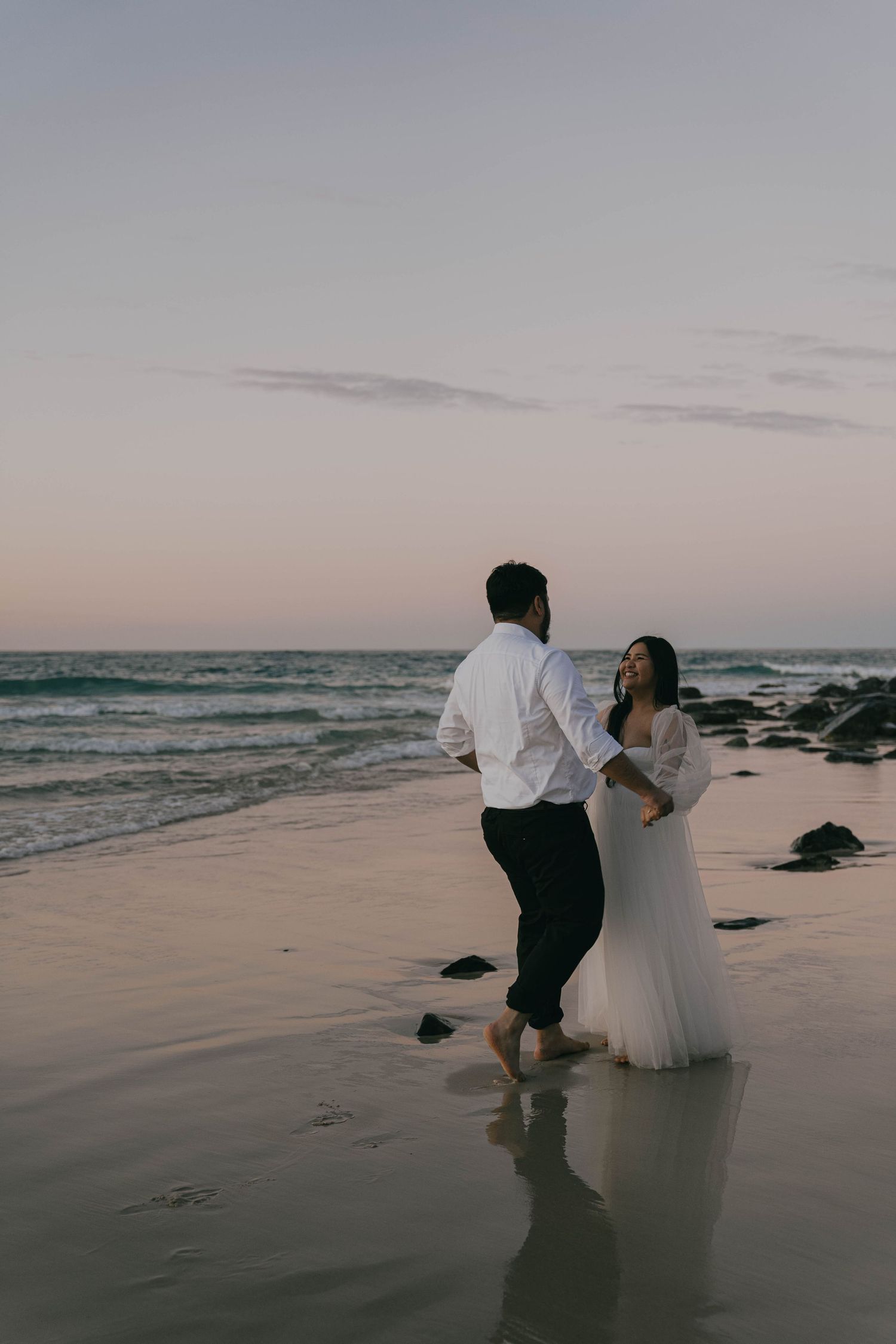 A couple in formal attire shares a romantic moment on a beach at sunset as waves crash on the shore.
