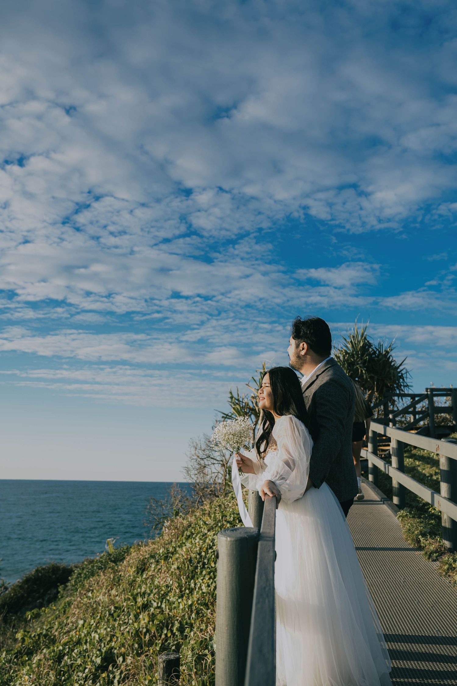 A couple in wedding attire stands on an oceanside boardwalk overlooking the sea under a dramatic blue cloudy sky.