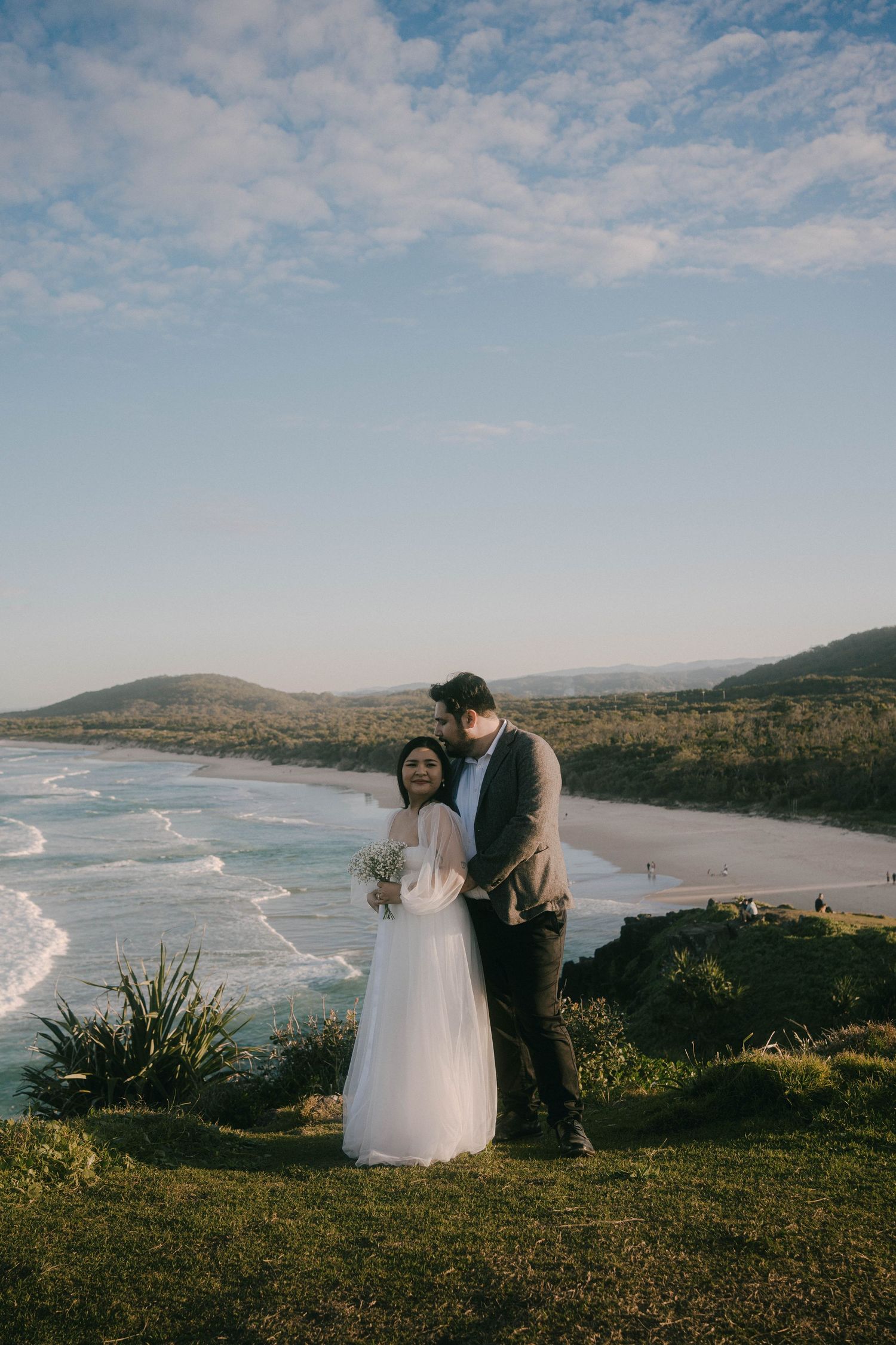 A couple in wedding attire stands on a scenic coastal hilltop overlooking a beach with rolling waves and mountains in the distance.