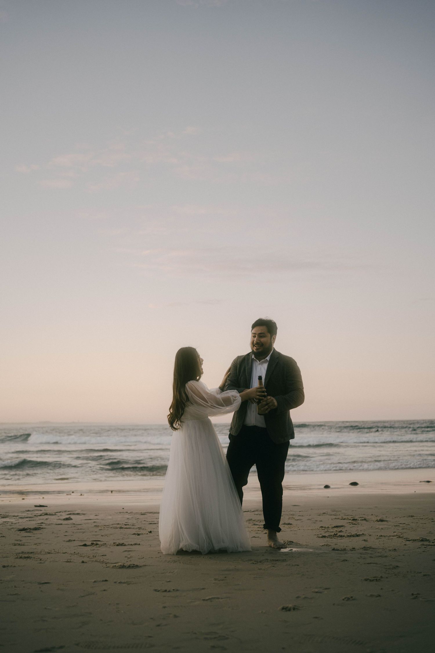 A couple in wedding attire walks along a misty beach at sunset, creating romantic silhouettes against the ocean waves.