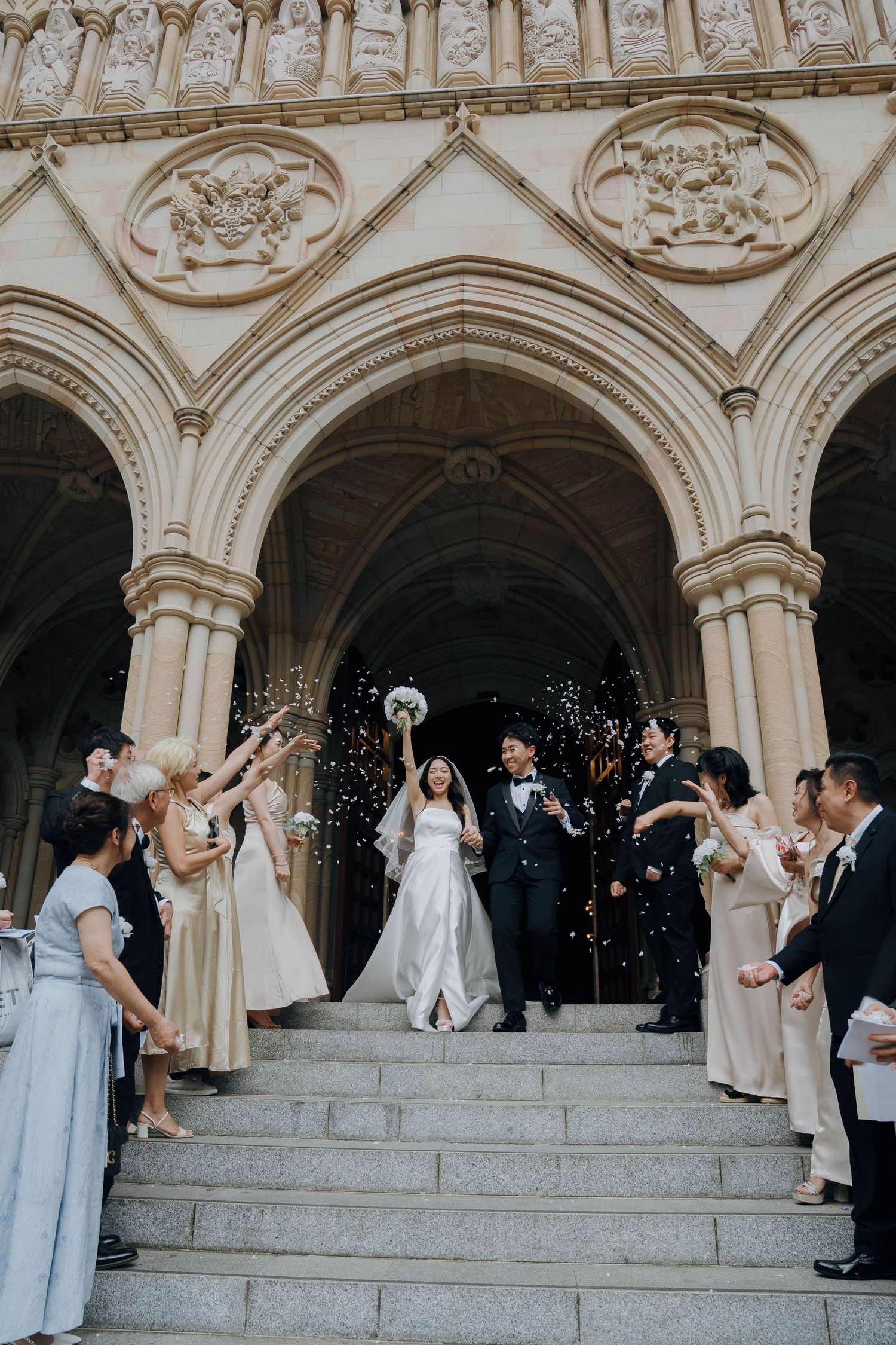 Wedding guests shower the newlyweds with confetti as they exit a grand cathedral with ornate Gothic arches and stone carvings.