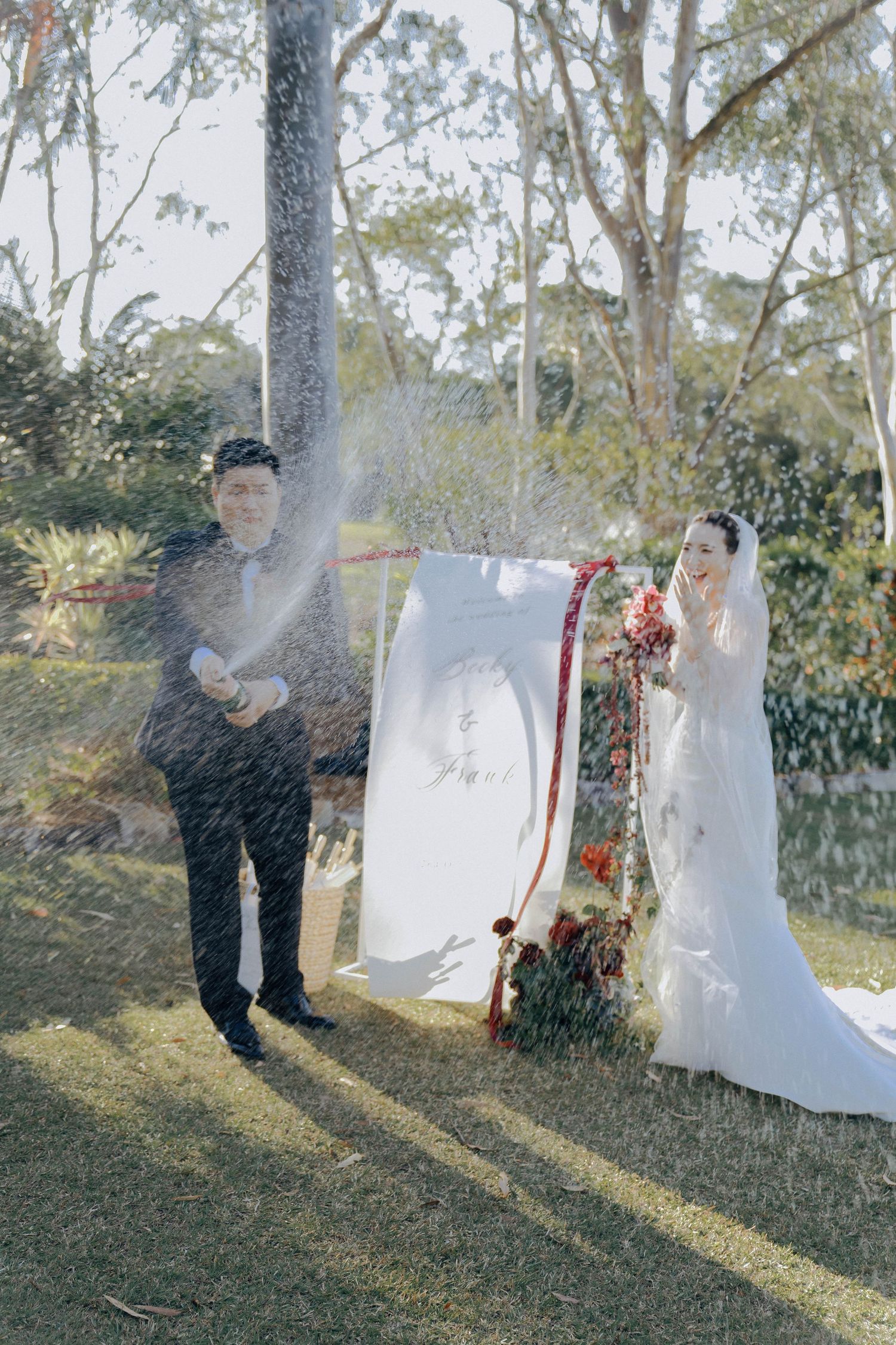 A couple poses outdoors with a white backdrop while a spray of water creates a dreamy effect in the sunlit garden setting.