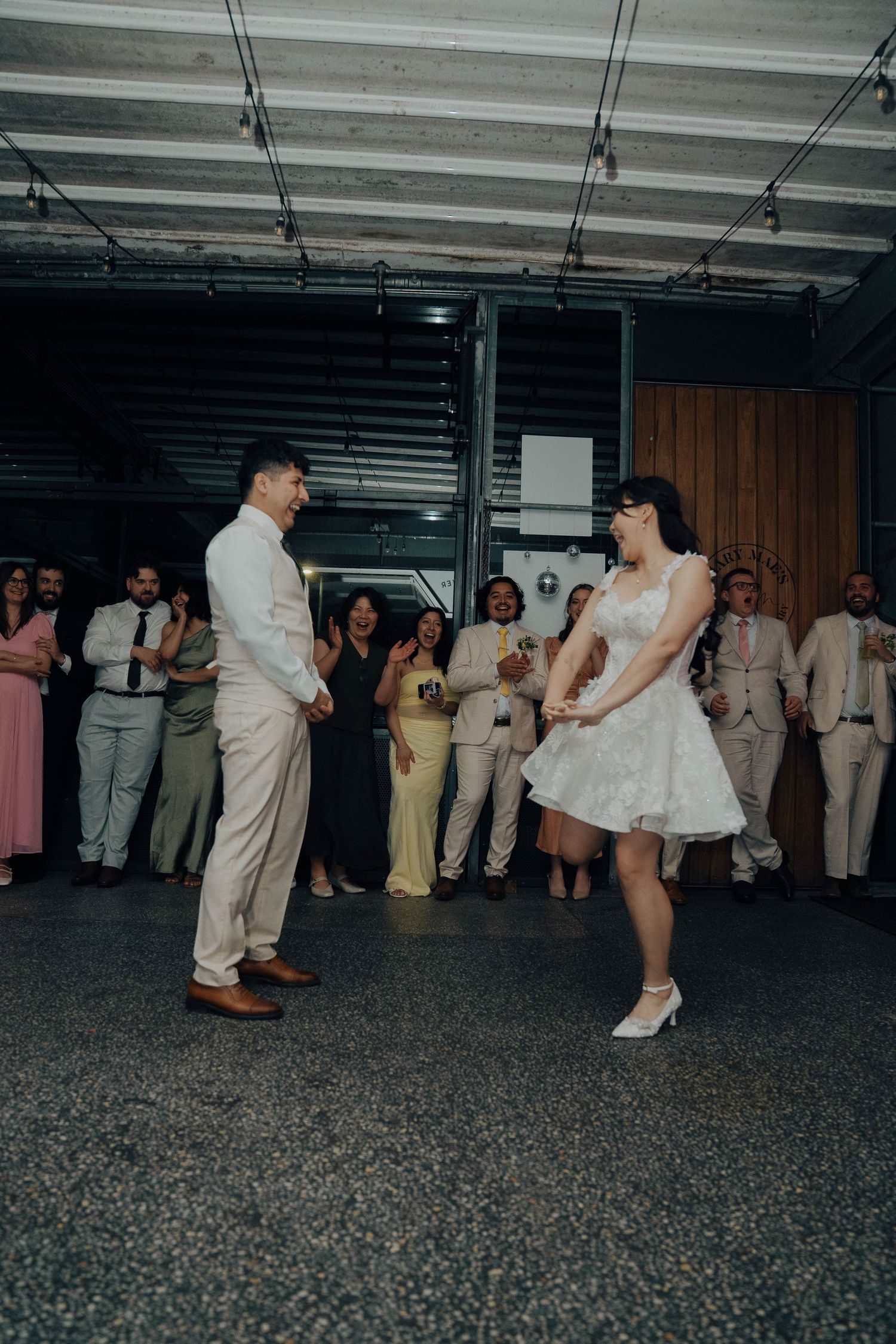 Wedding guests dance together on a dark dance floor at a reception celebration with people in white and pastel attire.