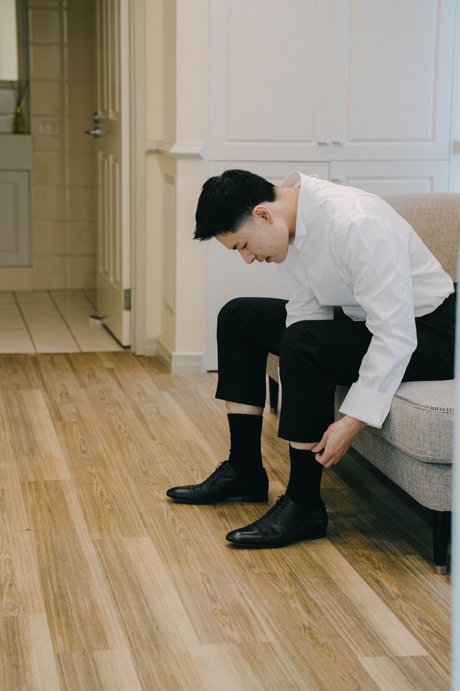 Person in white dress shirt and black pants sits on gray couch putting on formal black leather shoes.