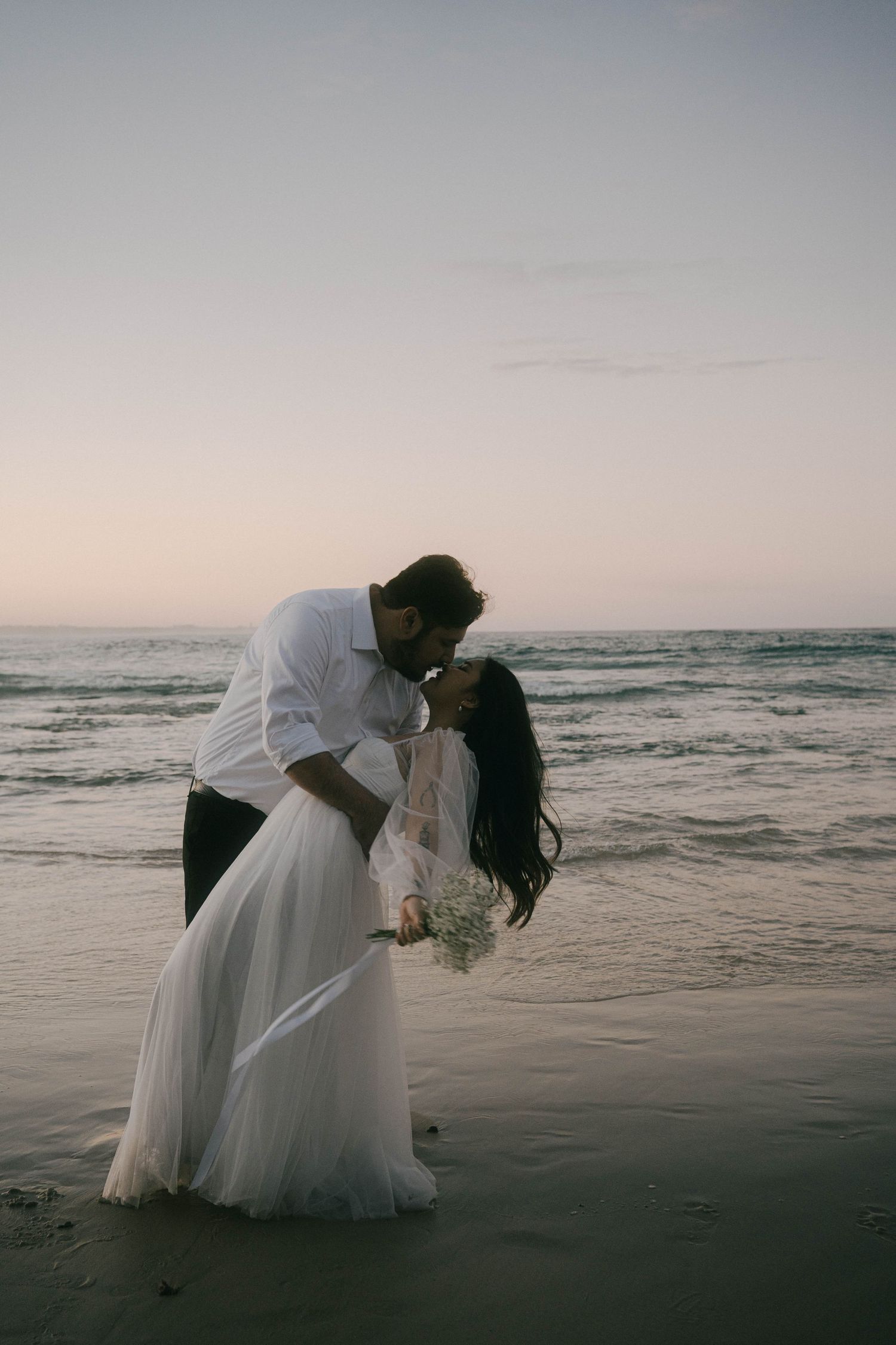 A couple in wedding attire shares an intimate moment on a moody beach at sunset.