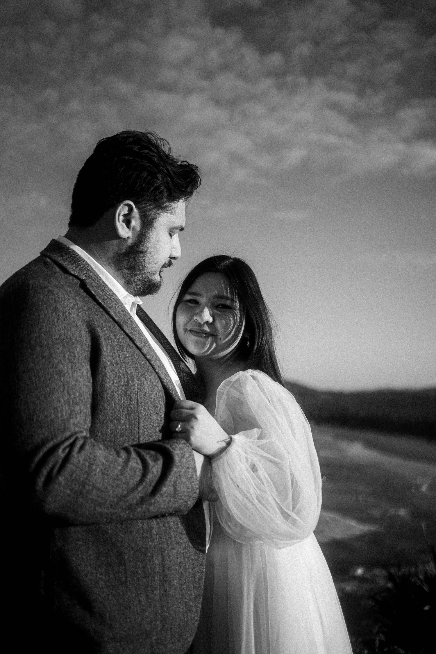 A romantic black and white portrait of a couple embracing outdoors with dramatic cloudy sky in the background.