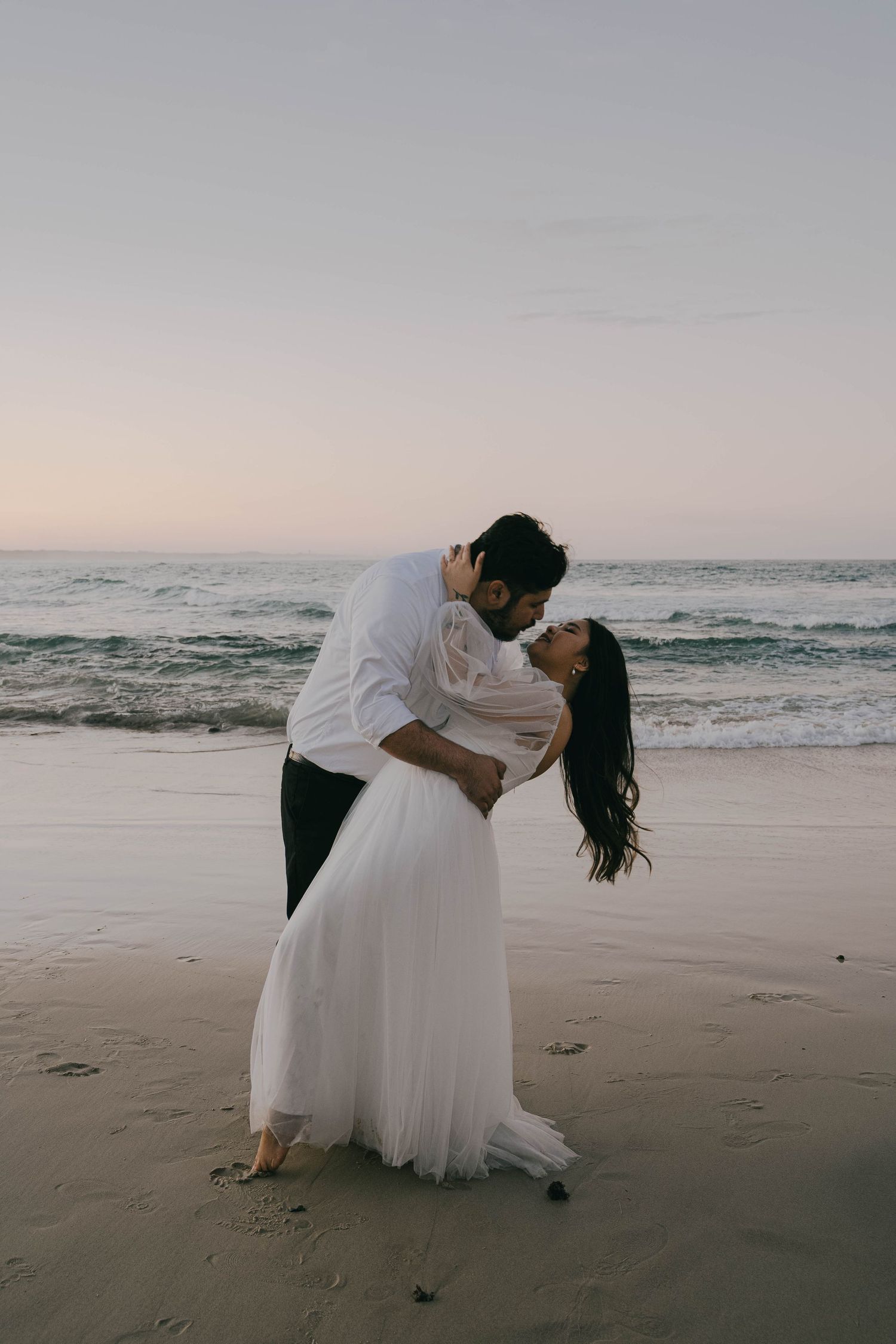 A romantic couple in white attire dances together on a misty beach at sunset.