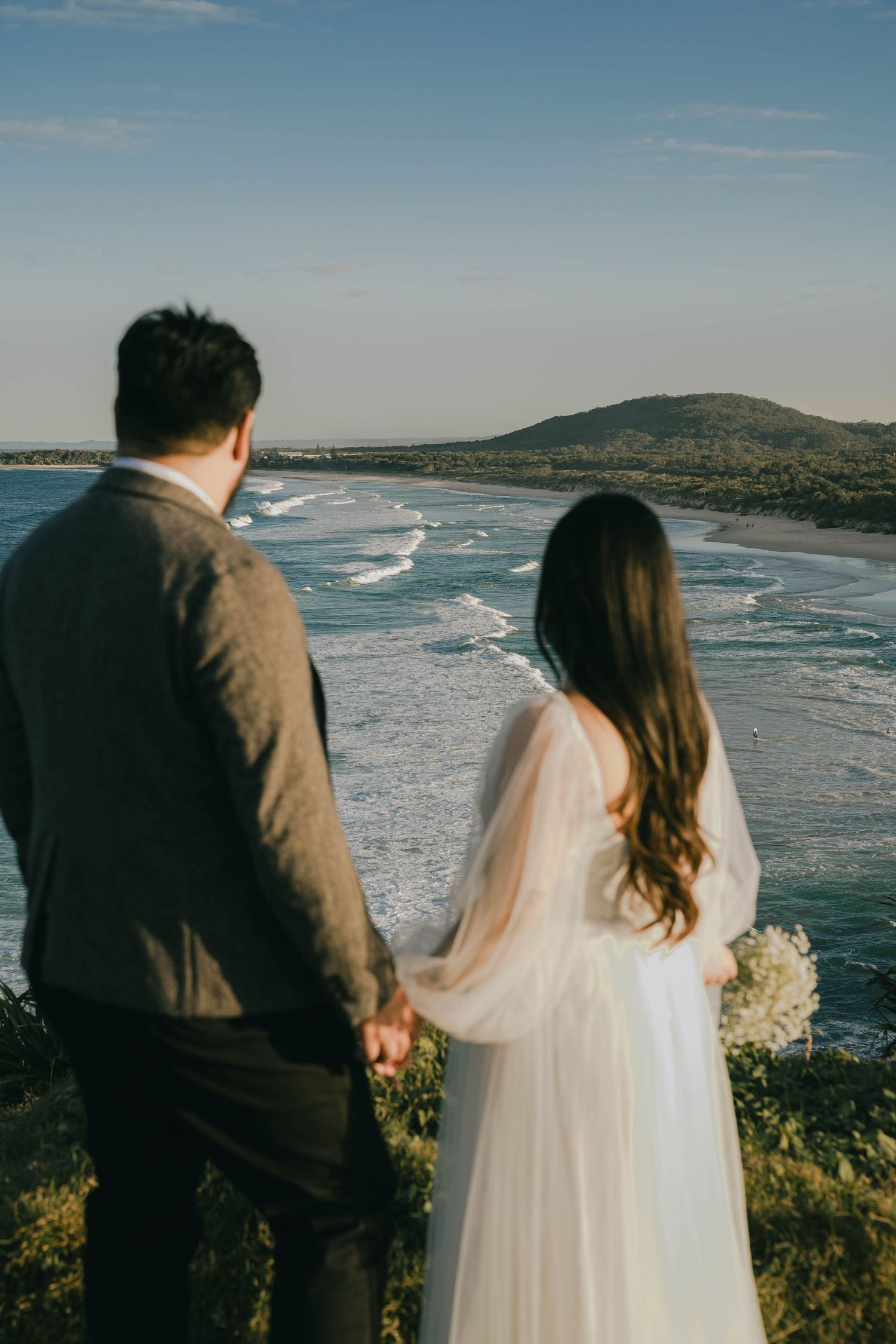 A couple stands together overlooking a scenic coastal landscape with crashing waves and distant mountains on their wedding day.