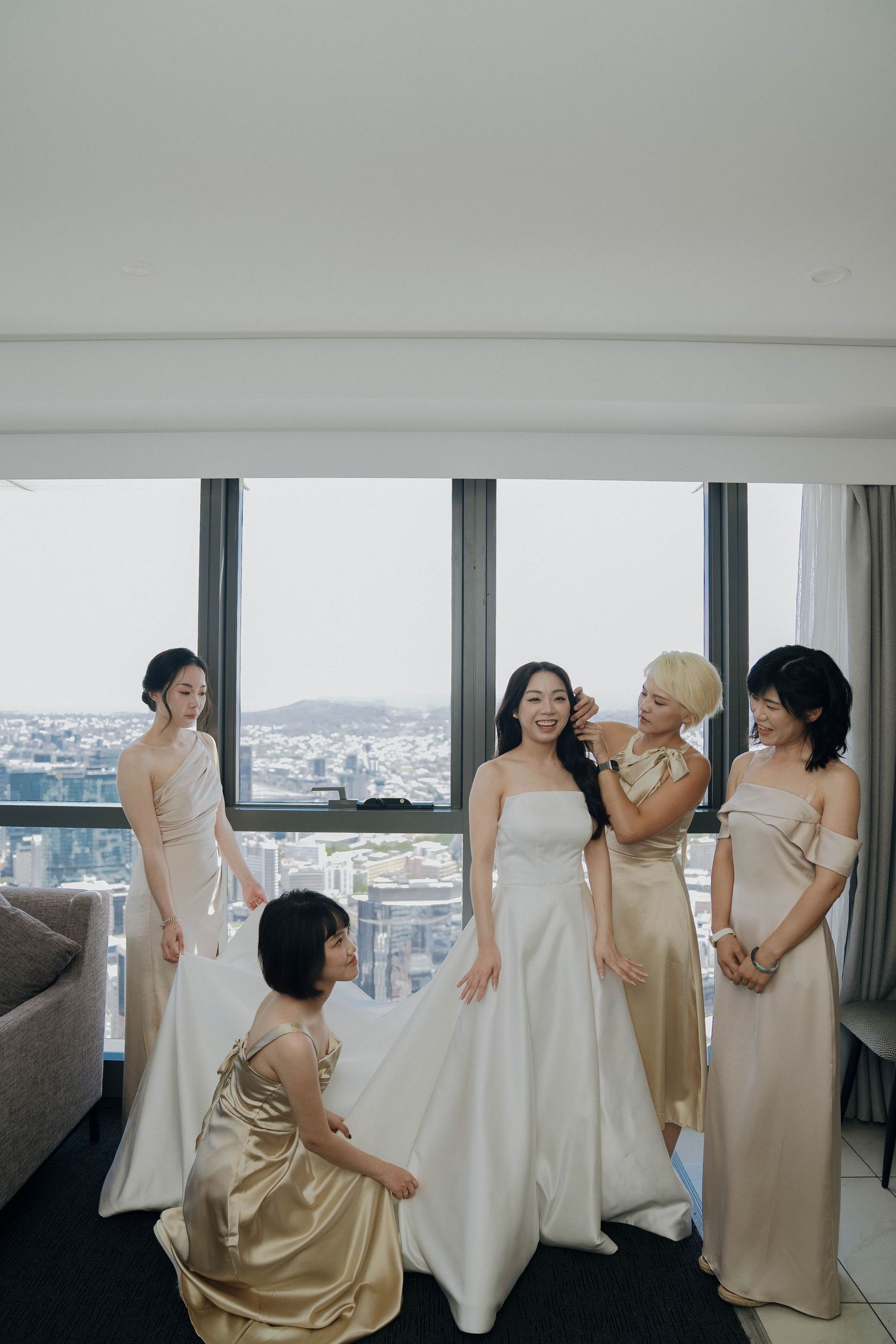 A group in elegant champagne and white formal dresses gather by large windows overlooking a cityscape on a cloudy day.