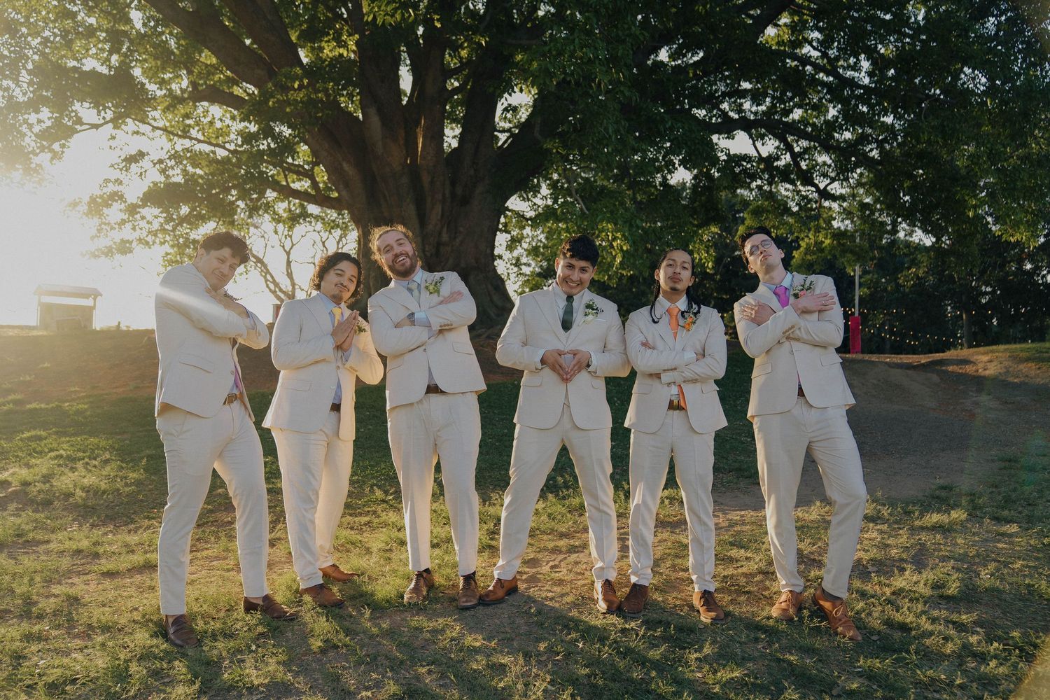 Wedding party members in coordinated white outfits stand together under a large tree at sunset.