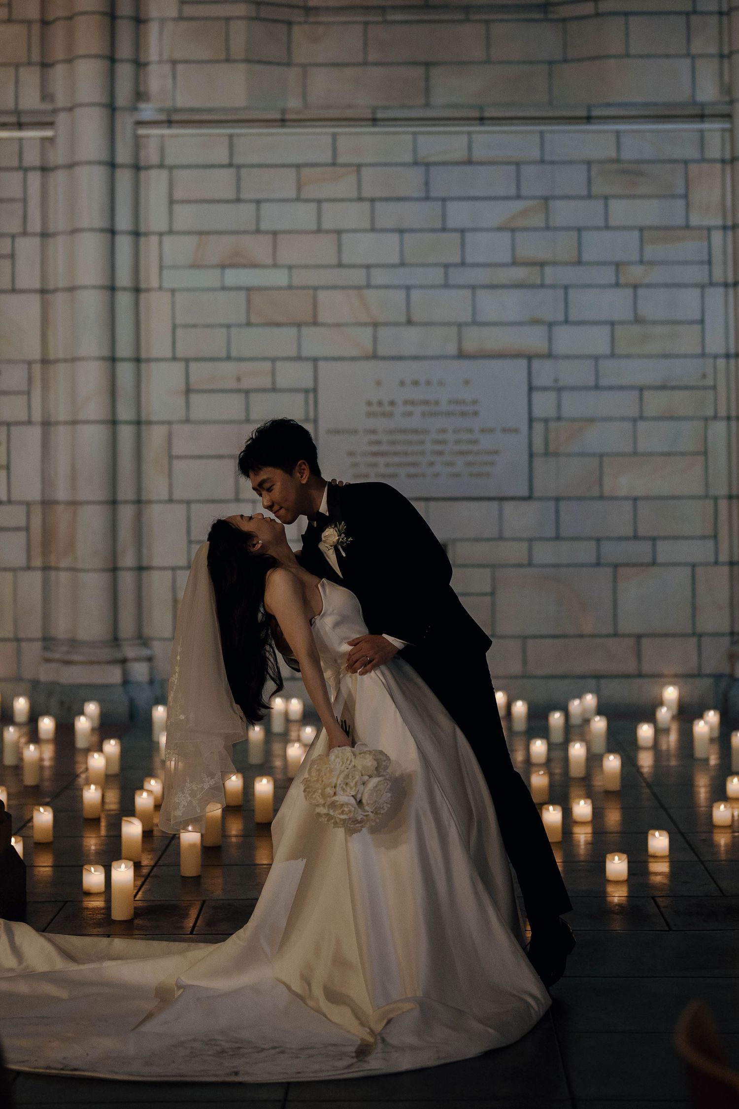 A romantic wedding couple shares an intimate kiss in front of glowing candlelight against a stone wall backdrop.