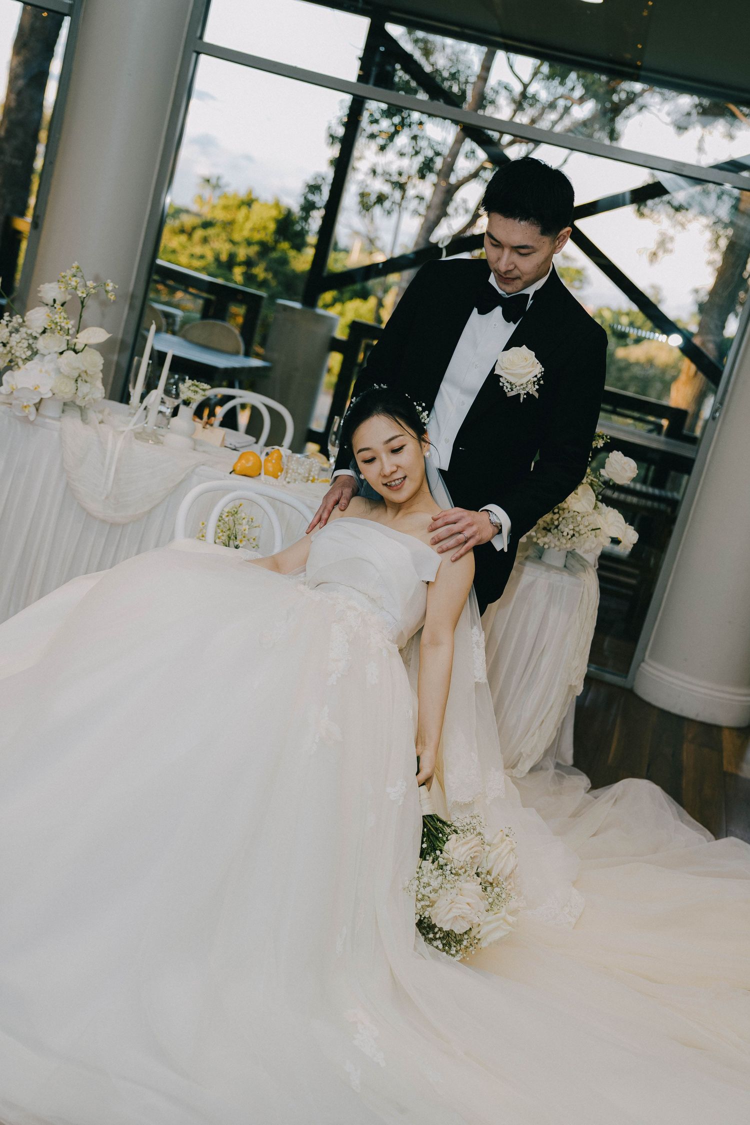 A couple in wedding attire poses together at an elegant venue with white floral decorations and natural lighting.