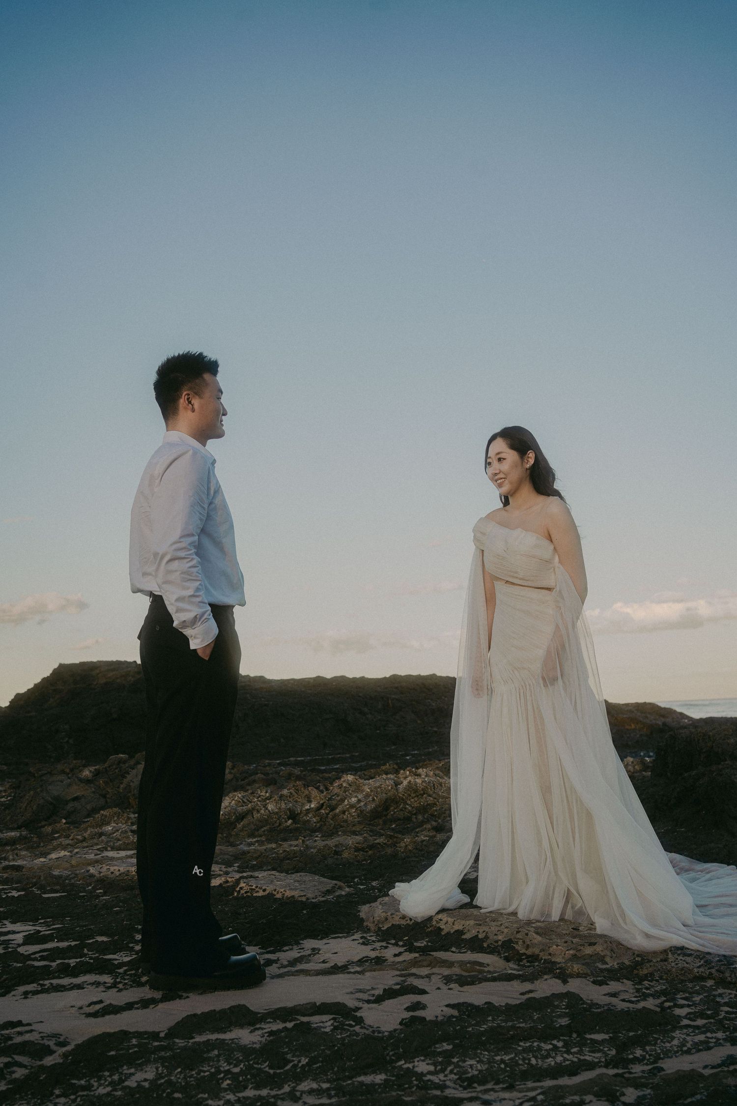 A couple stands on rocky coastal terrain at sunset, with the bride wearing a flowing white dress and groom in formal attire.
