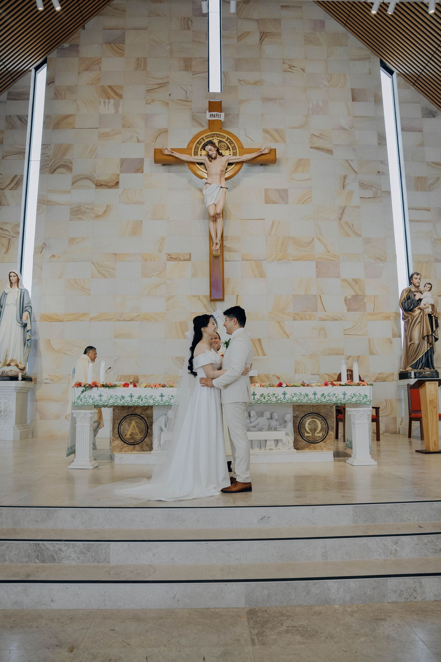 A couple stands at the altar during their wedding ceremony in a modern church with a large crucifix mounted on the wall.