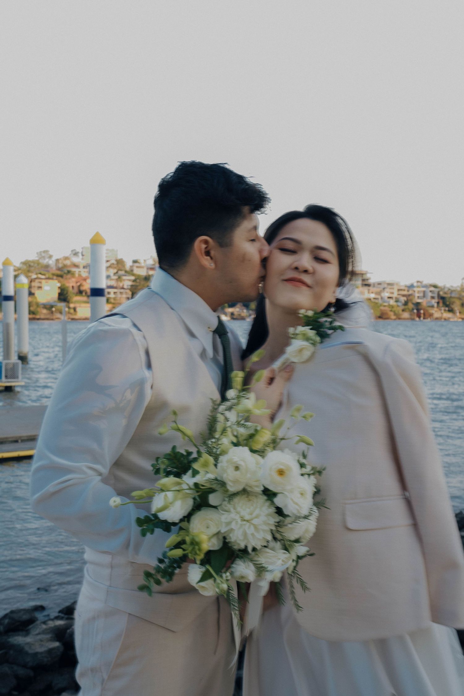 A romantic couple in matching white outfits share intimate moments by a waterfront during their wedding photoshoot.
