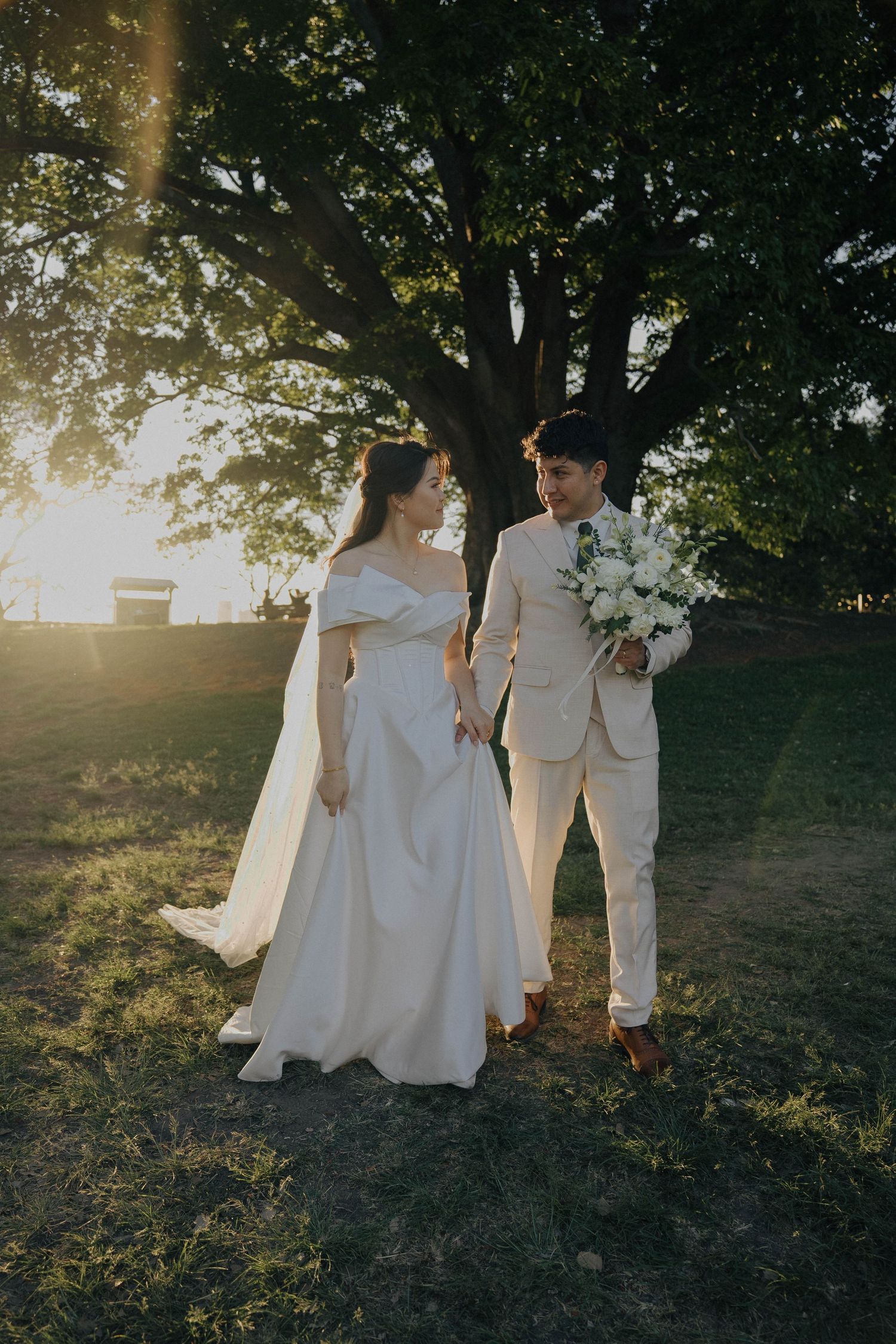 A romantic wedding couple stands under a large tree during golden hour, with the bride in white and groom in cream.