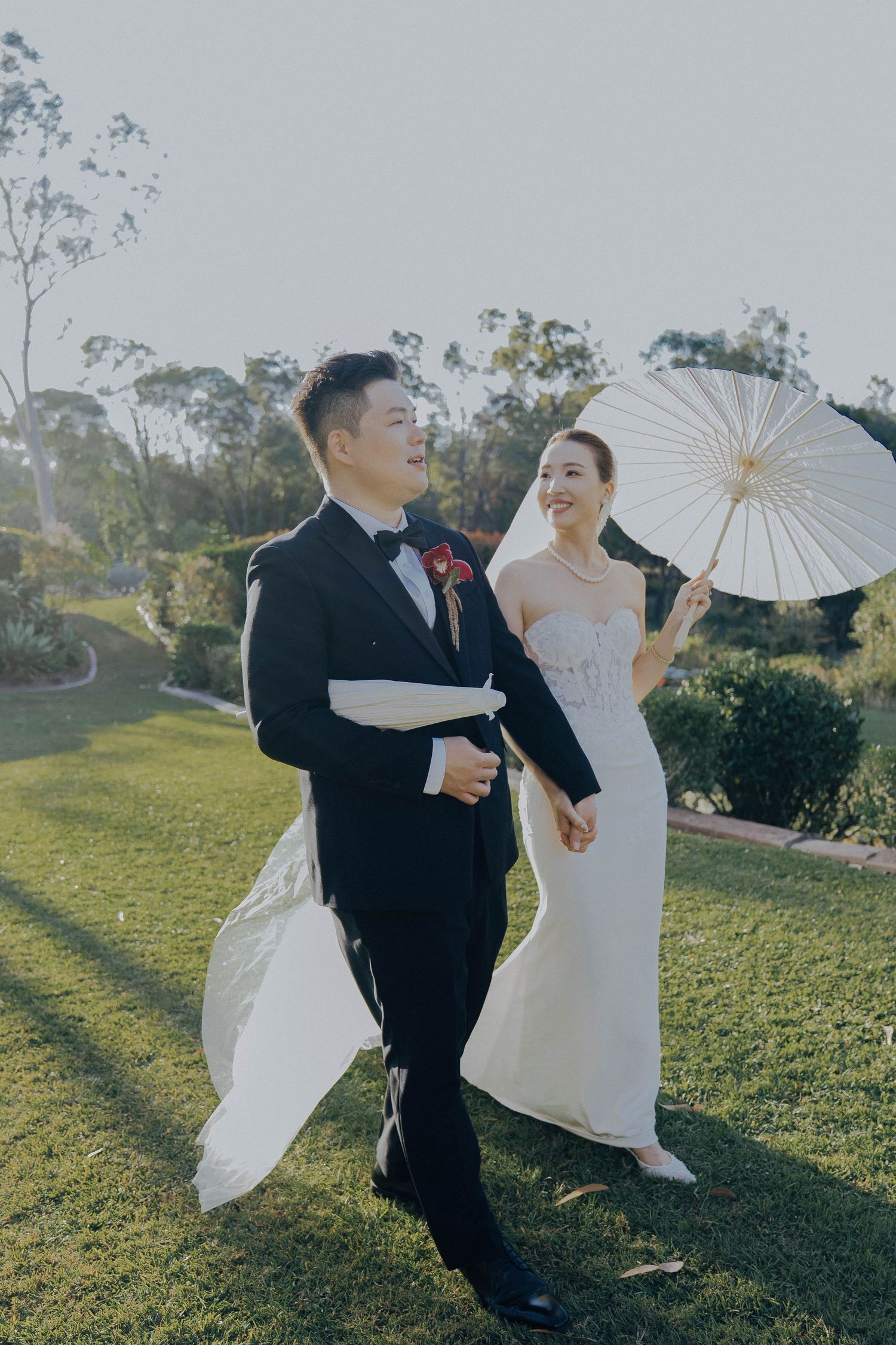 A couple in wedding attire dances playfully with white parasols in a sunlit garden setting.