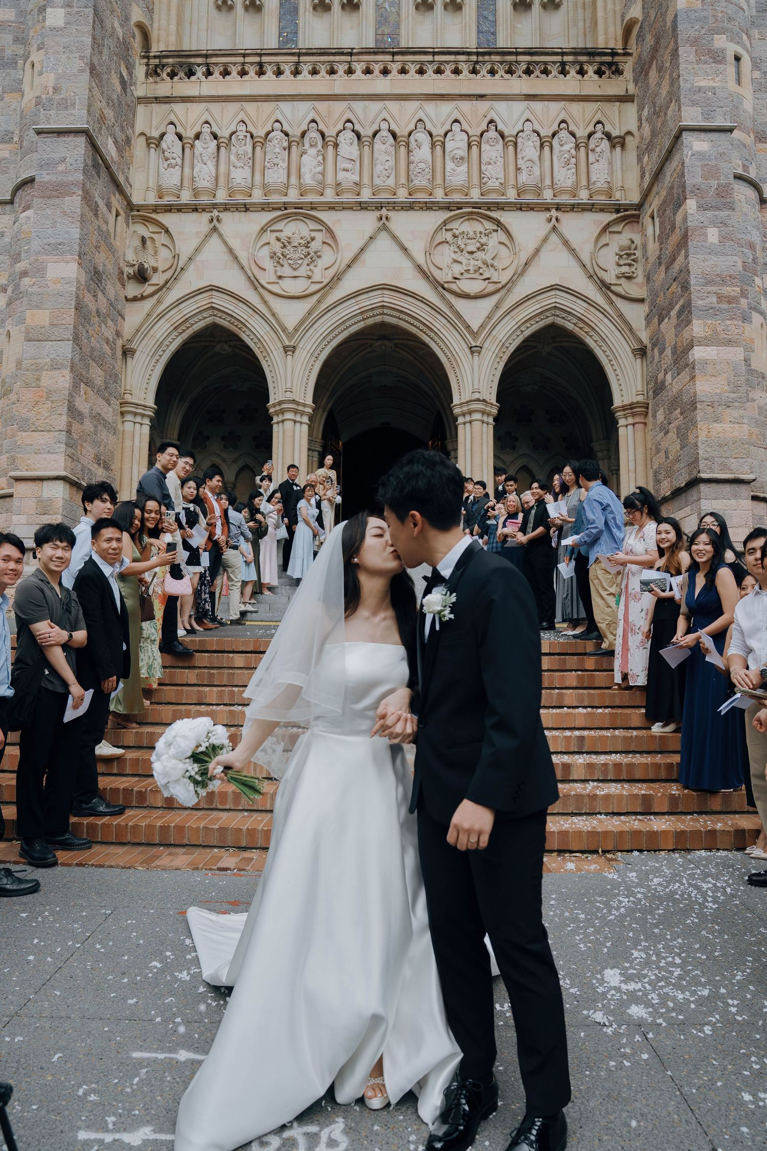 Newlyweds walk down church steps surrounded by guests throwing confetti in celebration of their wedding ceremony.