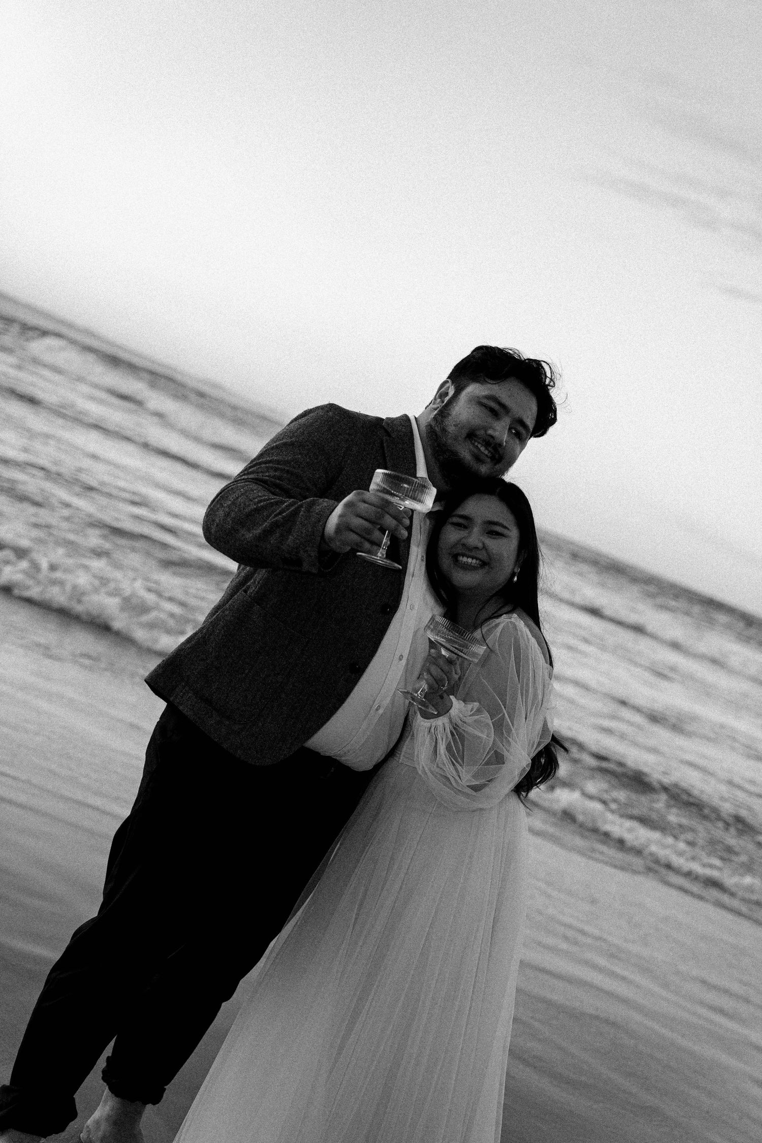 A couple shares a joyful moment together on the beach at sunset in black and white photography.