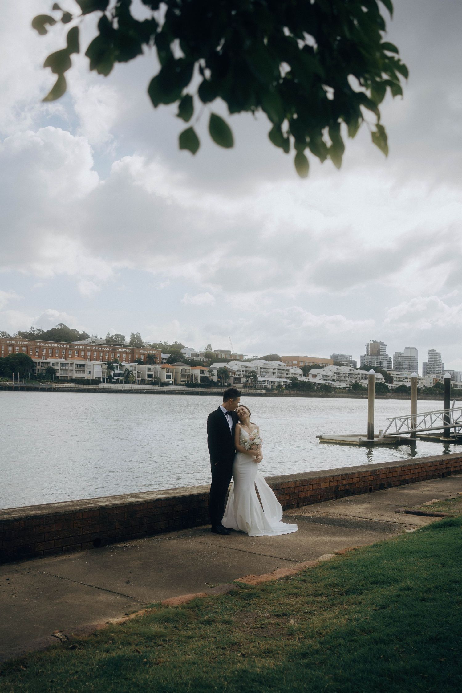 Newlywed couple shares romantic moments along a waterfront pier with city skyline views on an overcast day.