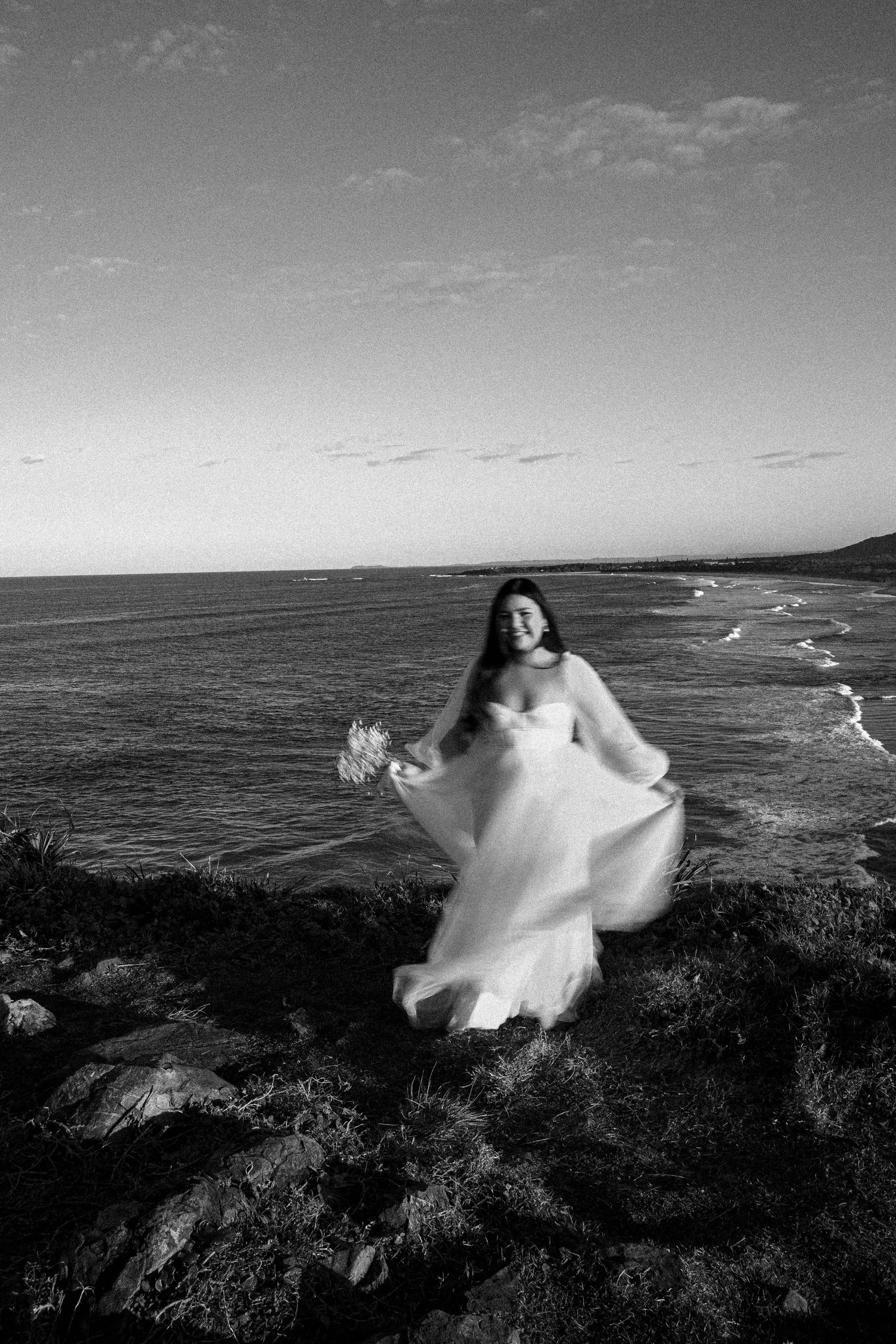 A bride in a flowing white dress twirls on a coastal cliff overlooking the ocean in dramatic black and white photography.