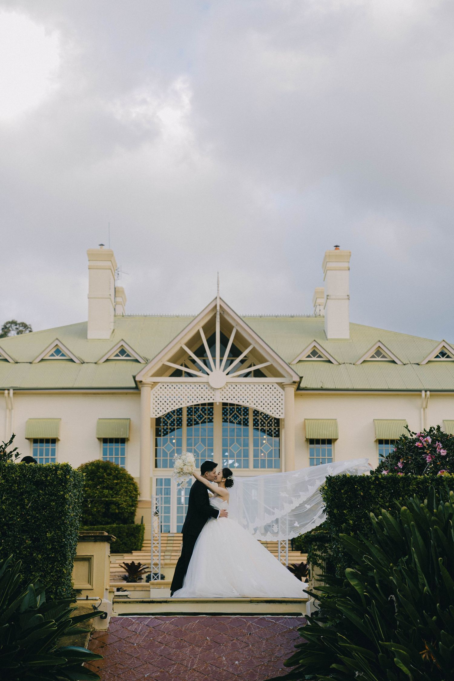A couple in wedding attire embrace in front of a historic cream-colored building with ornate architectural details.