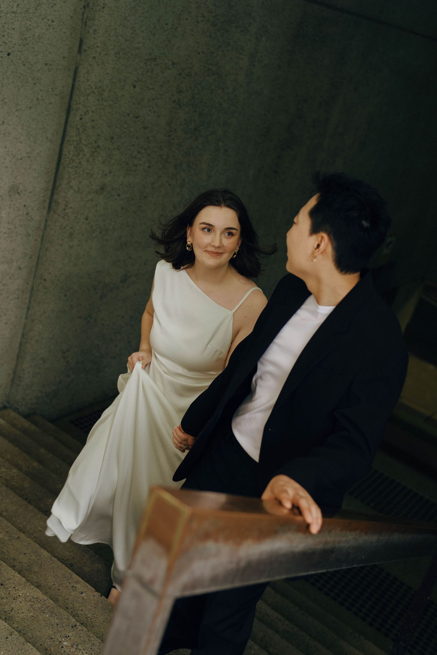 A couple in wedding attire sits together in a dark, moody setting with concrete walls.