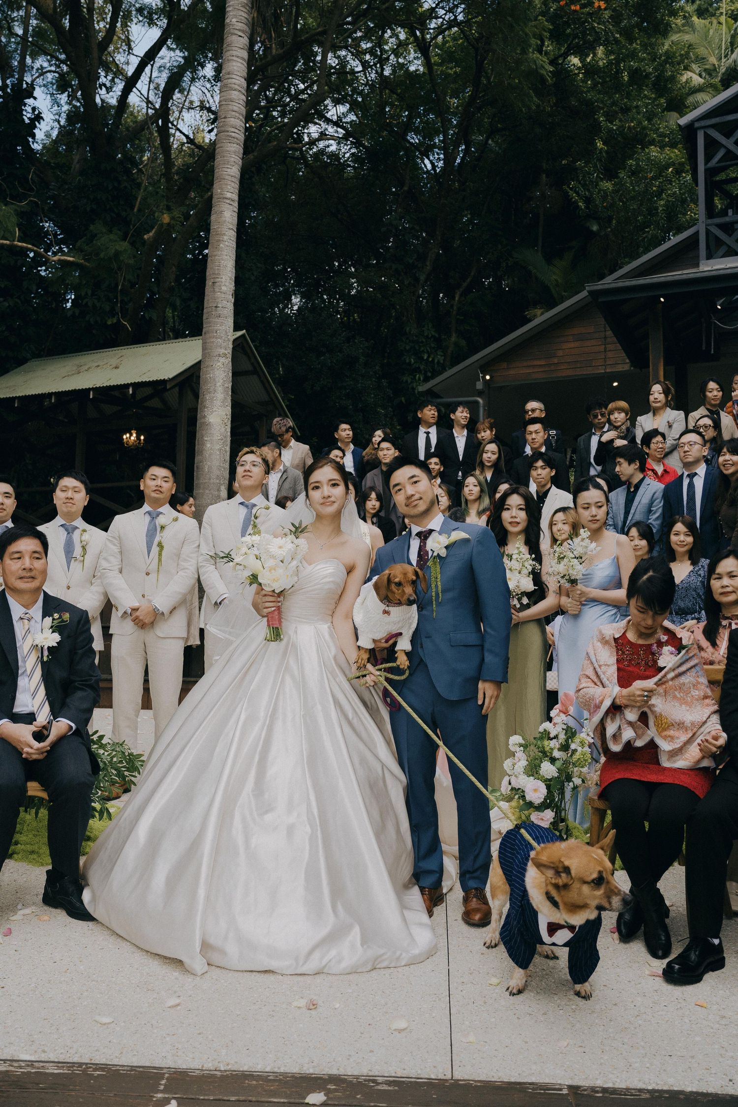 Large wedding party group photo captured outdoors with guests wearing navy suits and elegant white gowns.