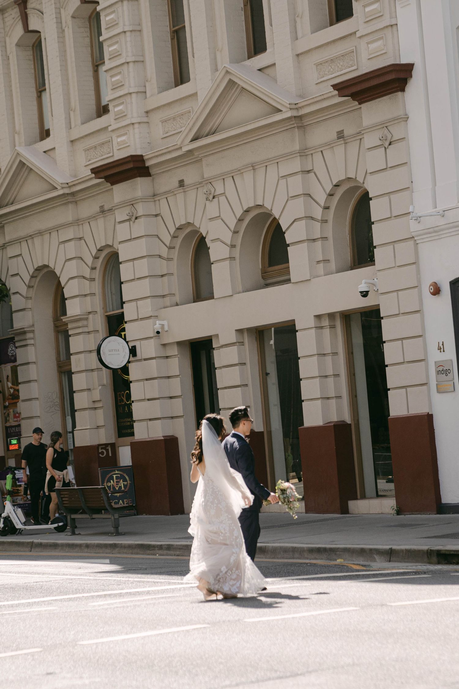 Couple in wedding attire walking through a grand archway with white stone pillars and classical architecture.