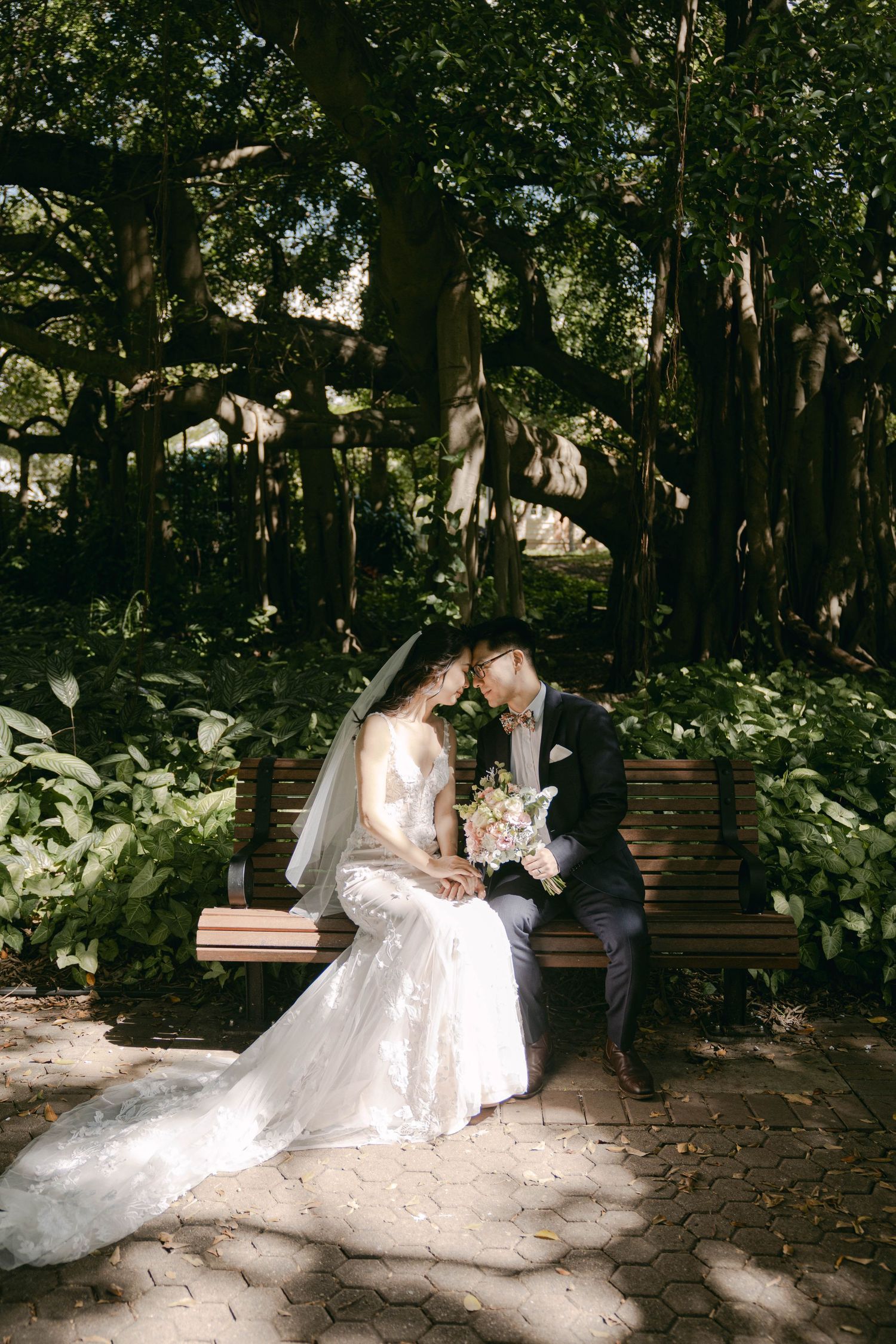 A couple in wedding attire share an intimate moment on a wooden bench beneath lush trees in a garden setting.