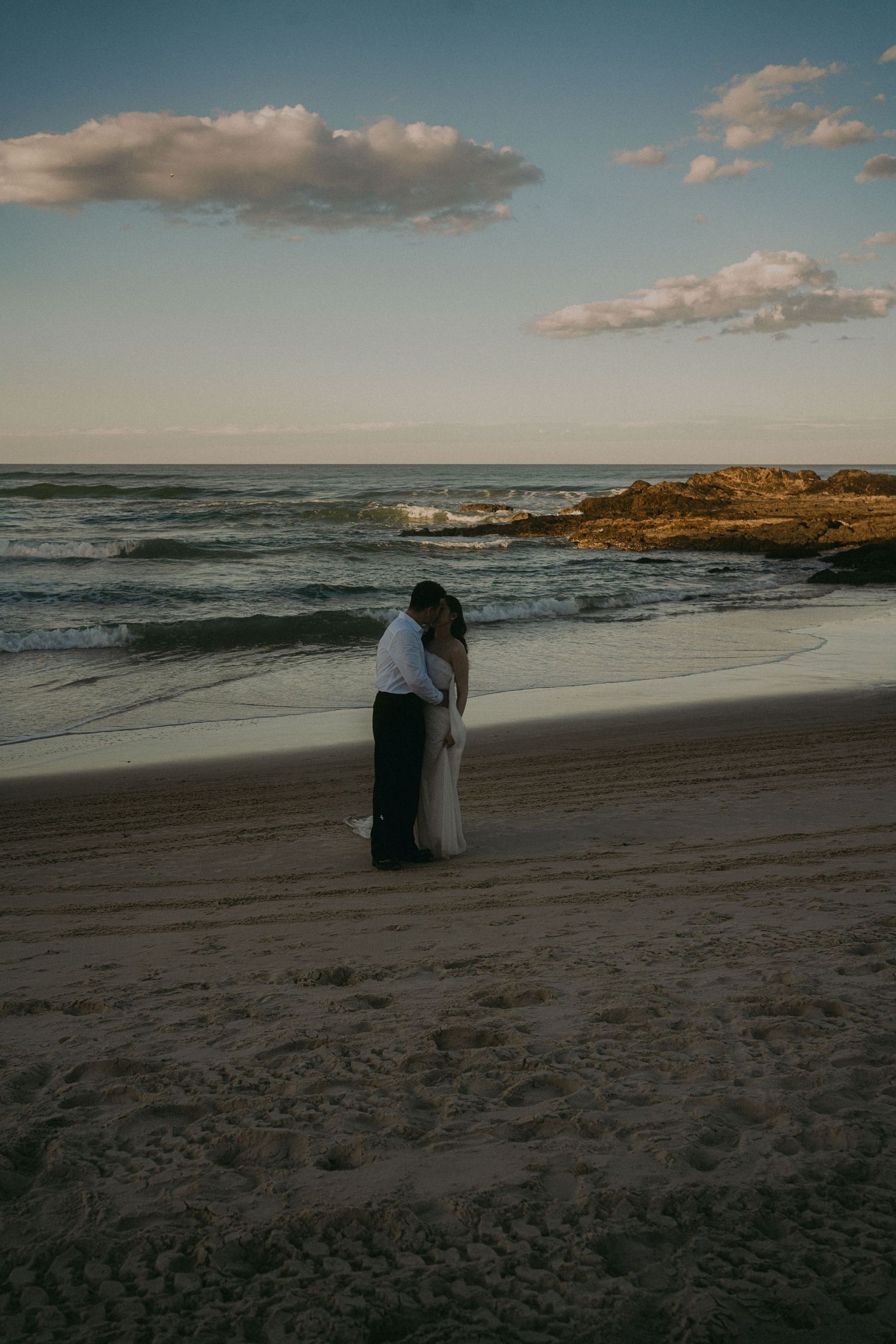 Two figures embrace while standing on a scenic beach at sunset with rocky coastline and rolling waves in the background.